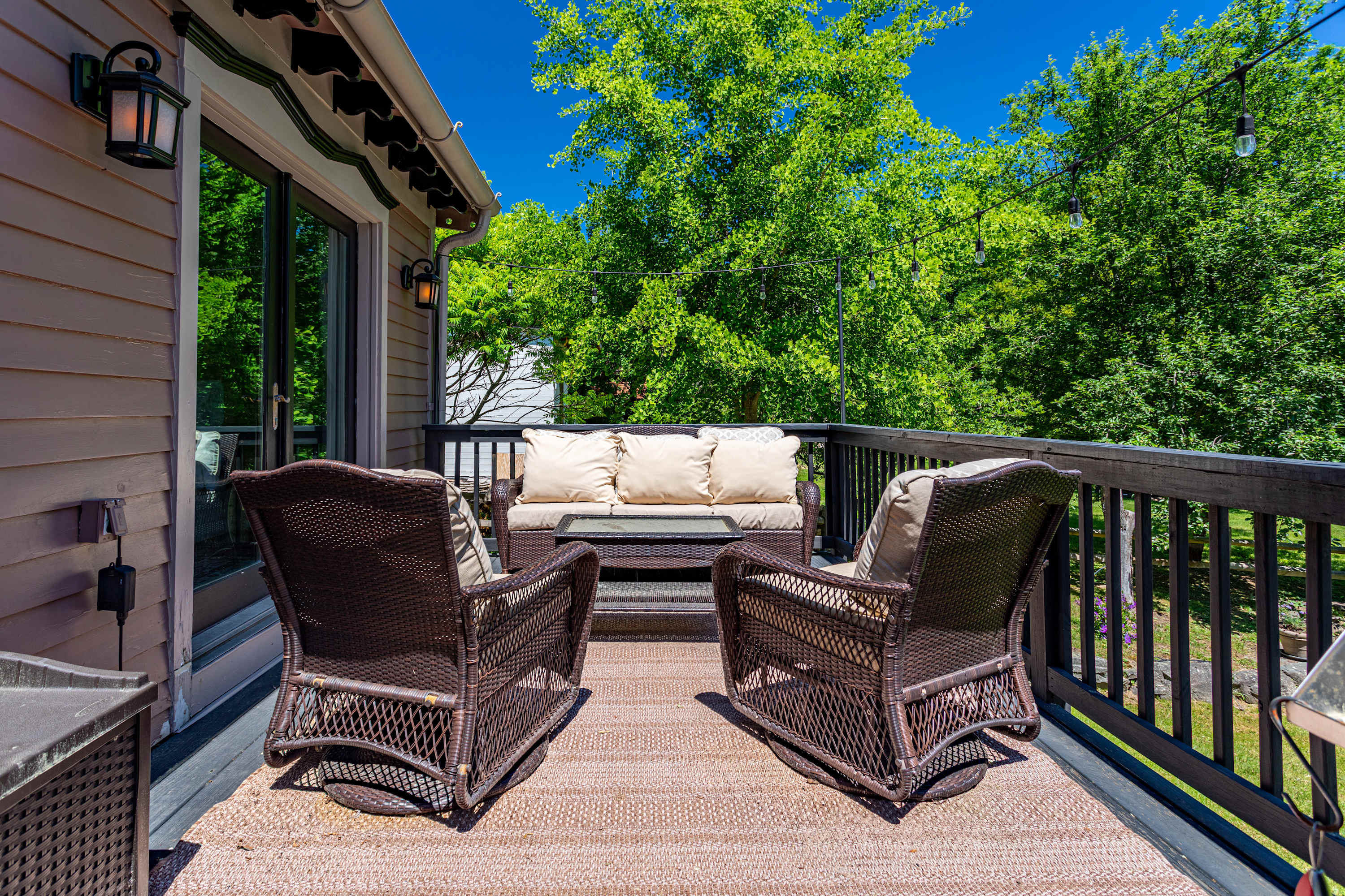 Cozy outdoor deck area featuring dark brown wicker lounge chairs and a matching sofa with light cushions, set on a woven rug, surrounded by a dark railing and lush green trees in the background.