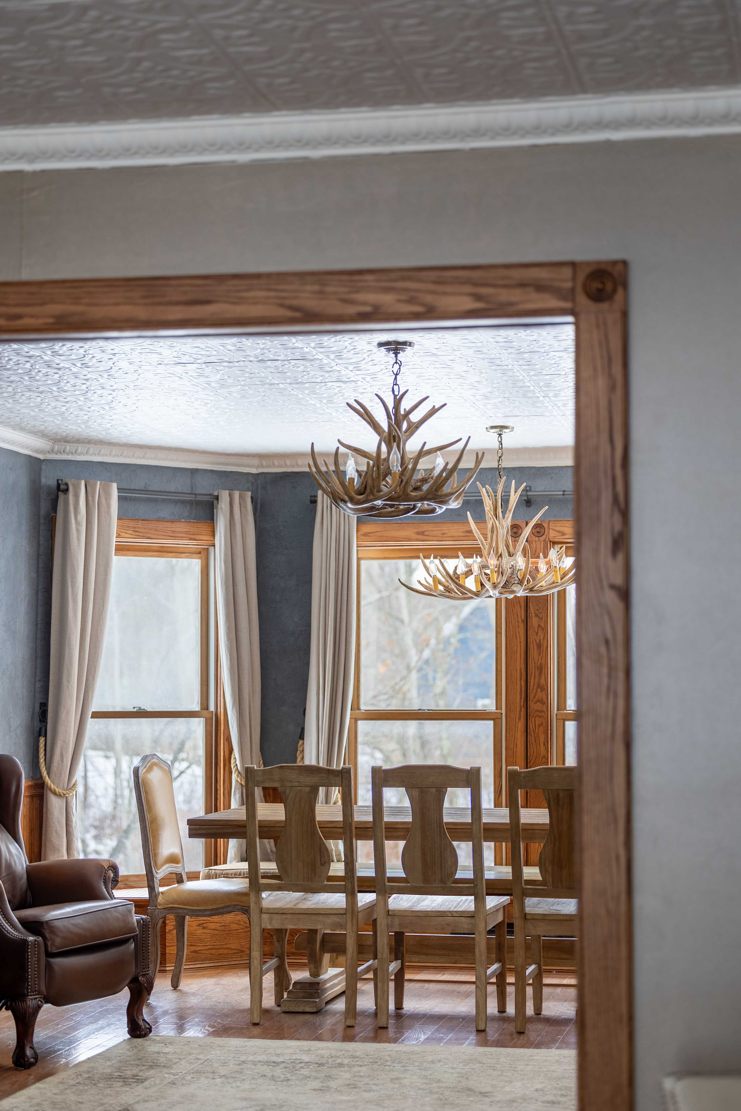 Elegant dining room with a large wooden table and matching chairs, illuminated by two rustic antler chandeliers, visible through a wooden doorway. The room features light-colored curtains and a decorative ceiling.