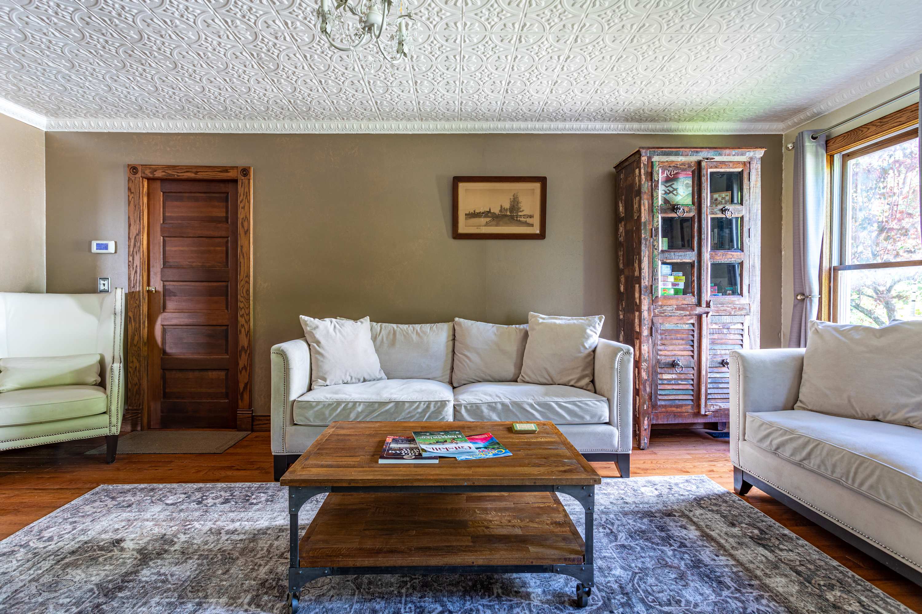 Cozy living room with a light gray sofa, wooden coffee table on a patterned rug, and a distressed wooden cabinet. The room features warm taupe walls, hardwood floors, and a white embossed ceiling.