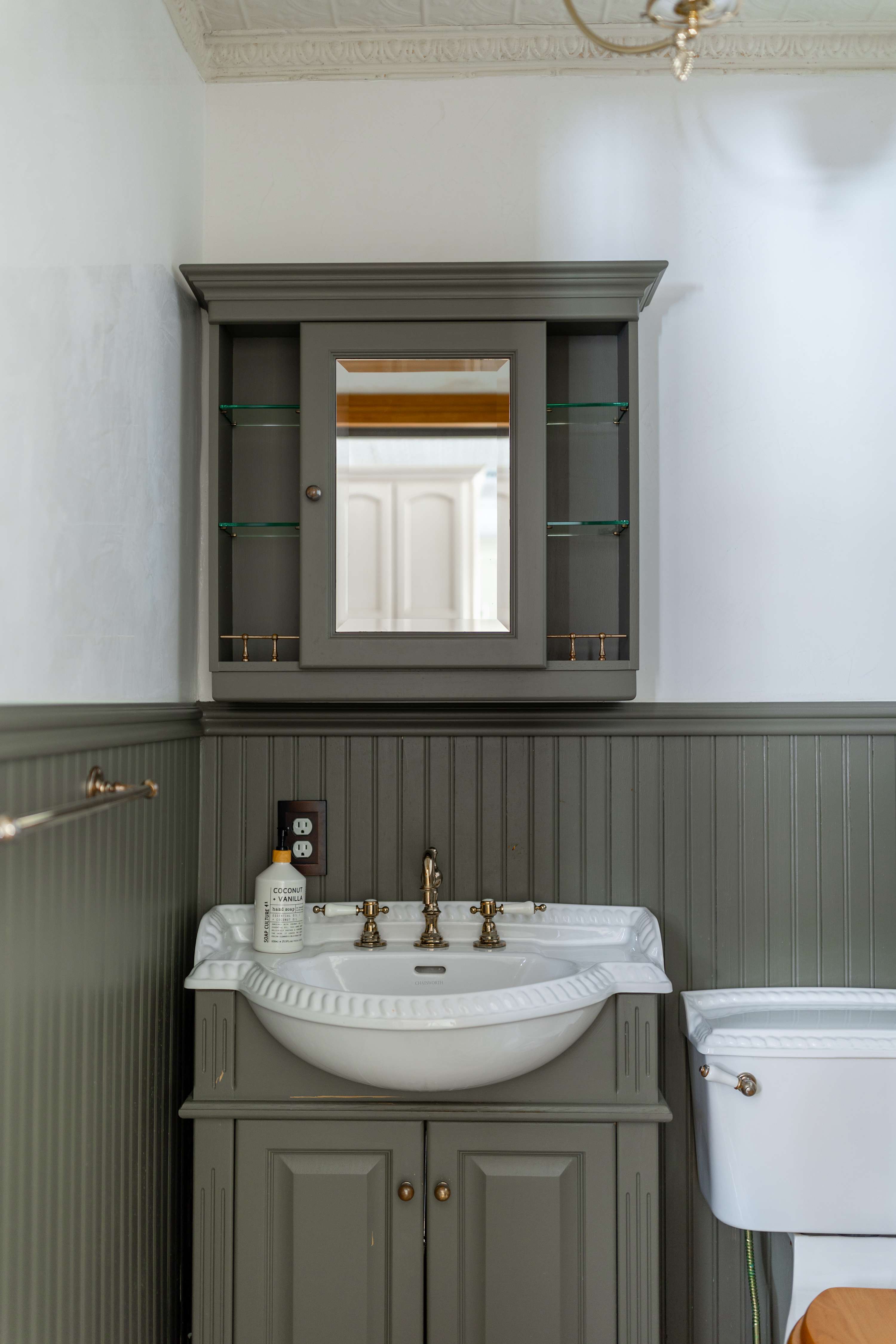 Classic bathroom featuring a gray vanity with a white basin sink and brass fixtures, a matching medicine cabinet with a mirror, and beadboard wainscoting. A white toilet is partially visible.