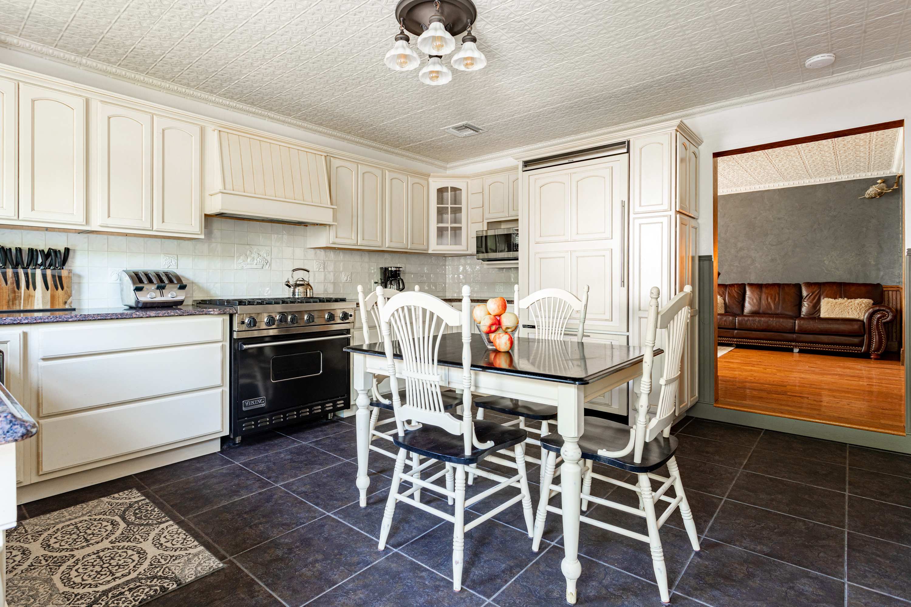 Spacious kitchen featuring white shaker cabinets, dark tiled floors, and a black Viking range. A central dining table with white chairs is set with fruit, and a built-in refrigerator is visible.