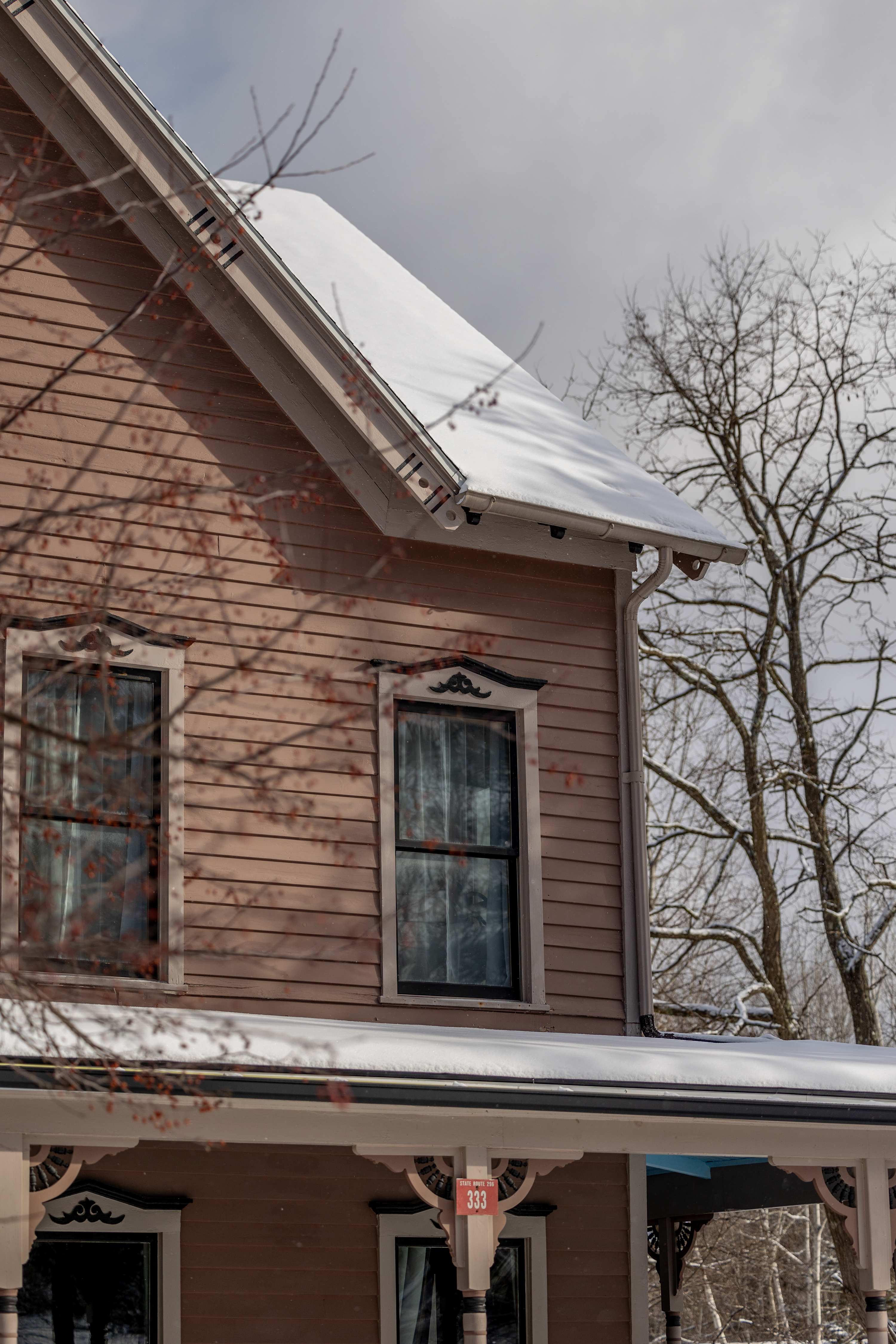 Side view of a multi-story house with light brown siding and dark trim, featuring snow-covered roofs and decorative gables. Bare trees stand against a cloudy sky, indicating winter.