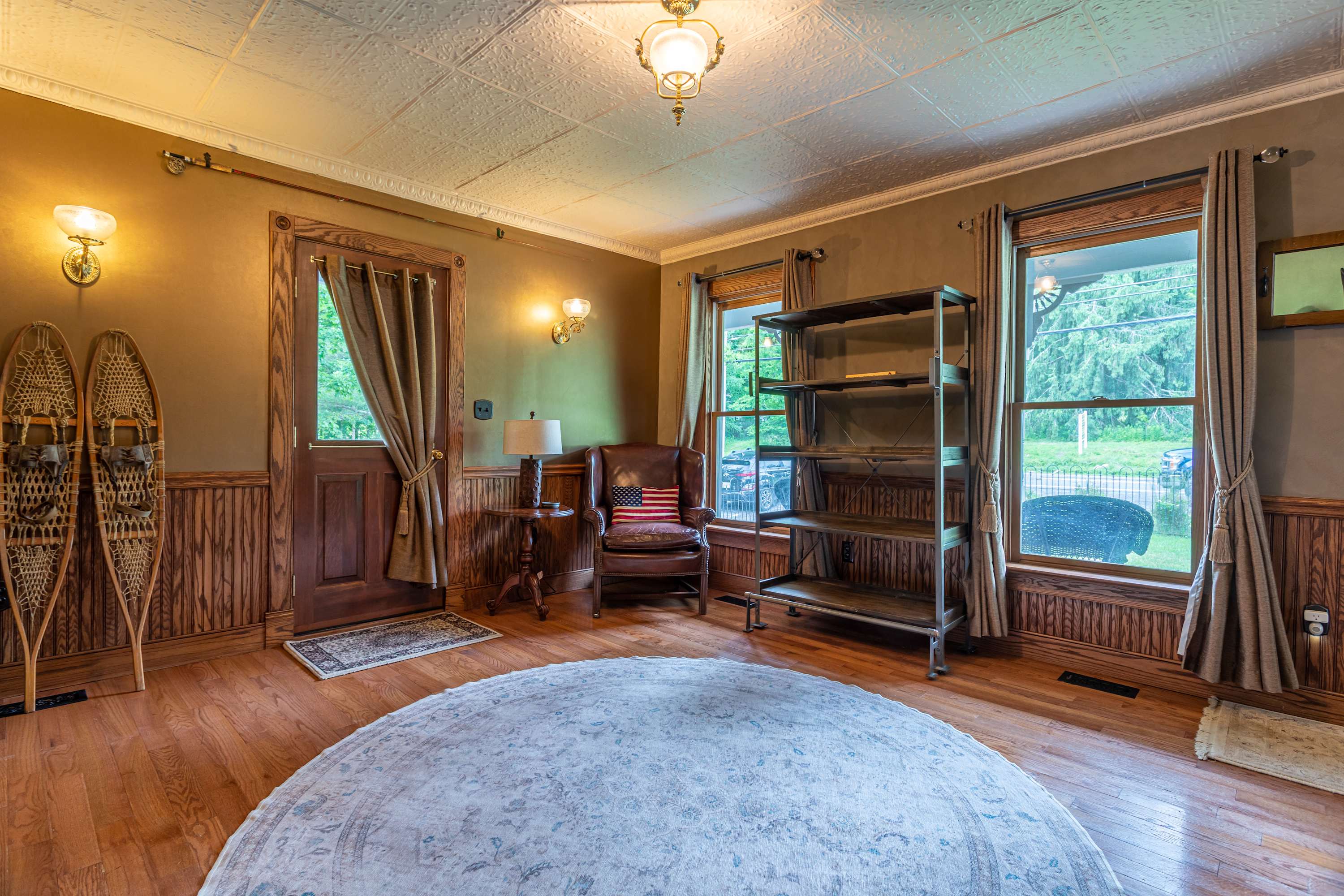 Rustic sitting area with a brown leather armchair, industrial-style bookshelf, and decorative snowshoes mounted on the wall. The room features warm taupe walls, wooden wainscoting, and hardwood floors.