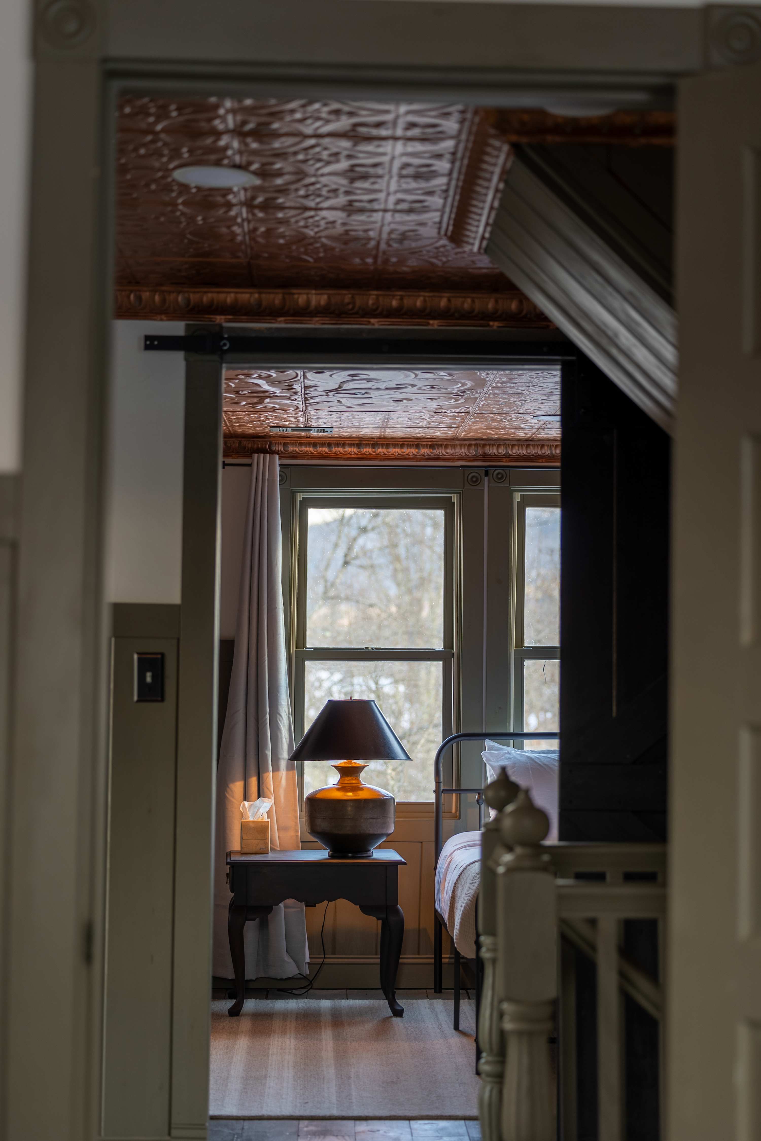 View into a bedroom through a doorway, revealing a bed with a metal frame, a dark nightstand with a lamp, and windows with light curtains. The room features a textured ceiling and warm lighting.