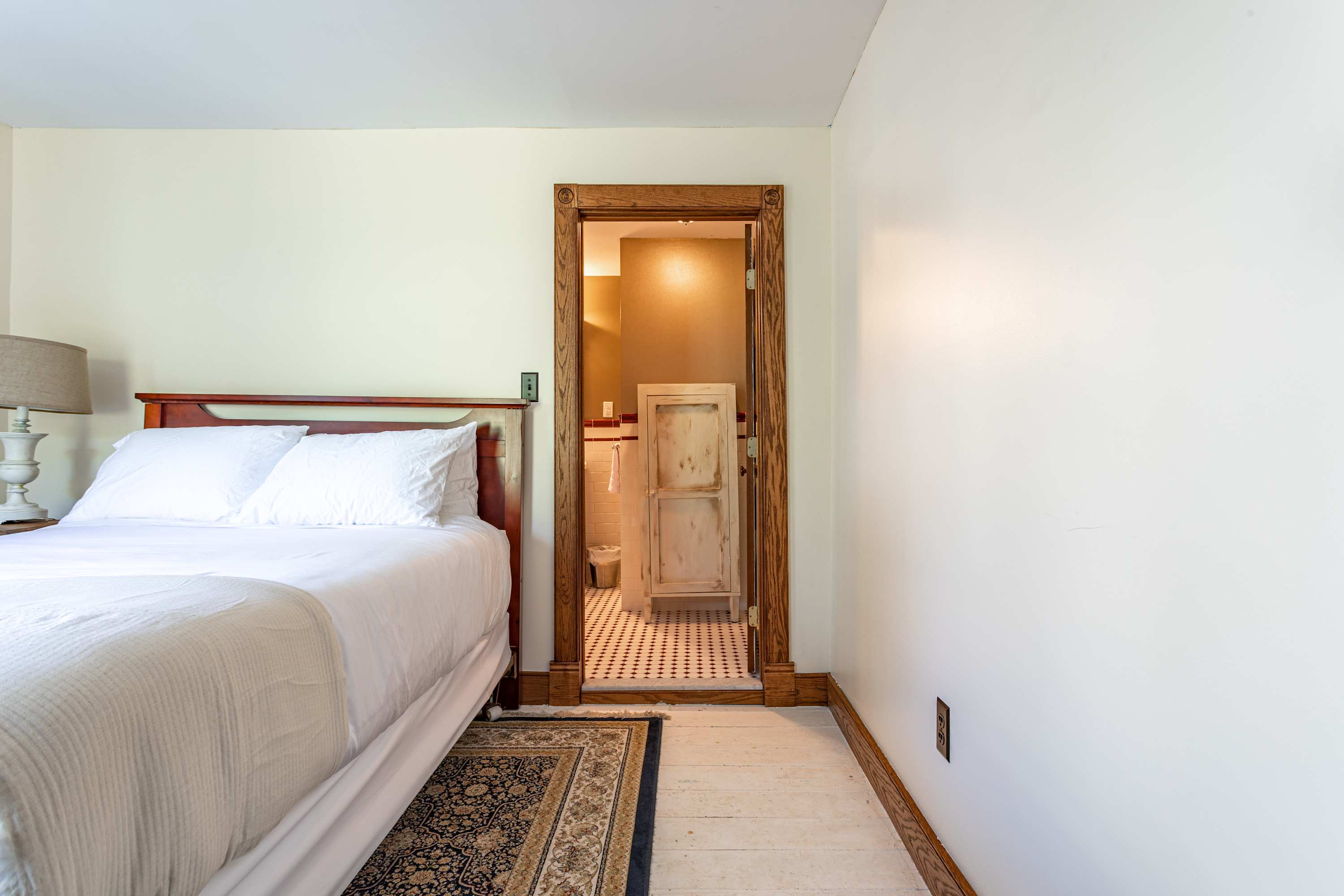 Bedroom with a wooden bed frame and white bedding, showing an open doorway leading into an en-suite bathroom with patterned floor tiles.