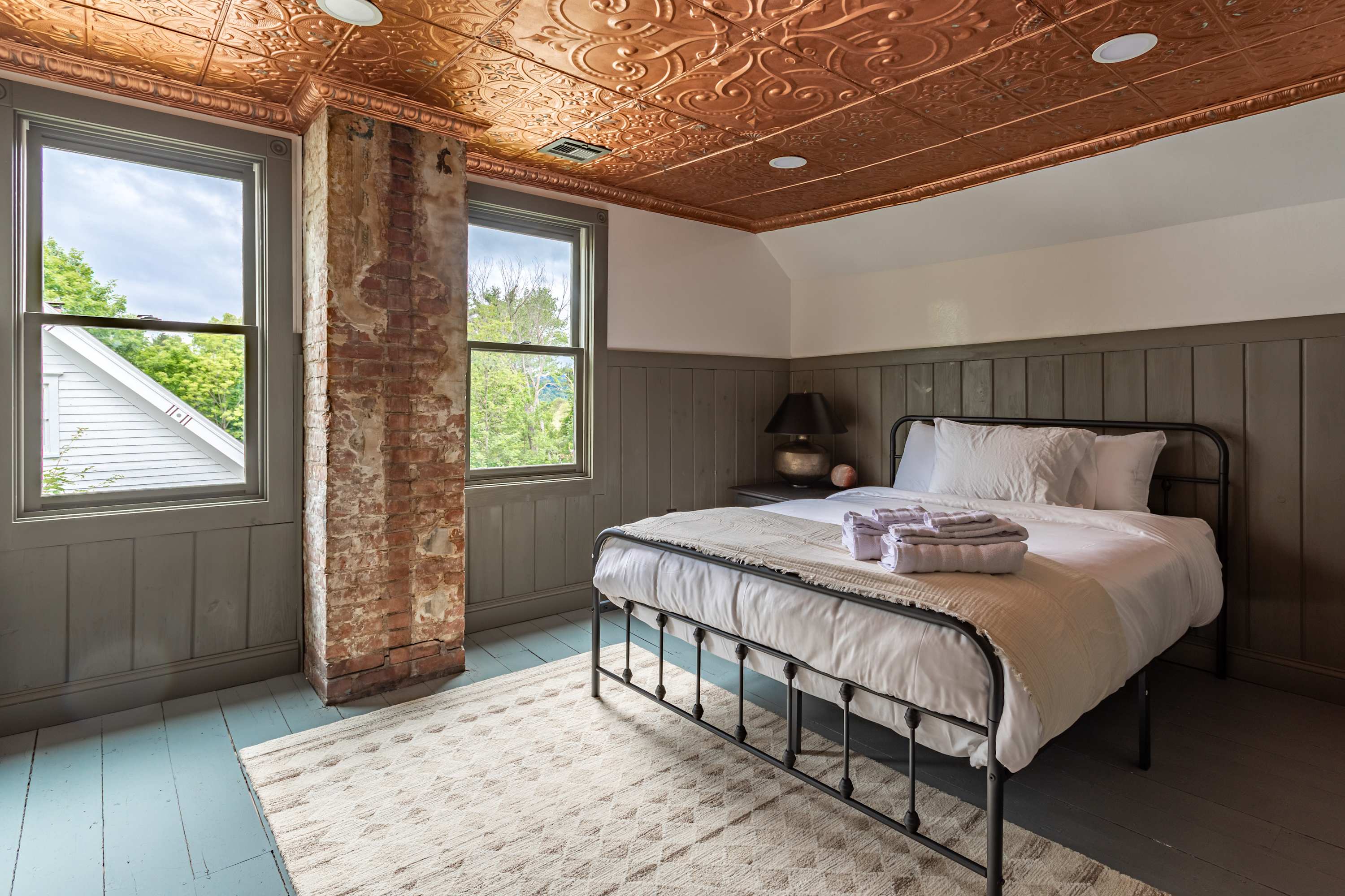 Bright bedroom with a black metal frame queen bed, an exposed brick column, light wood paneling, and two large windows, all beneath a copper-colored pressed tin ceiling.