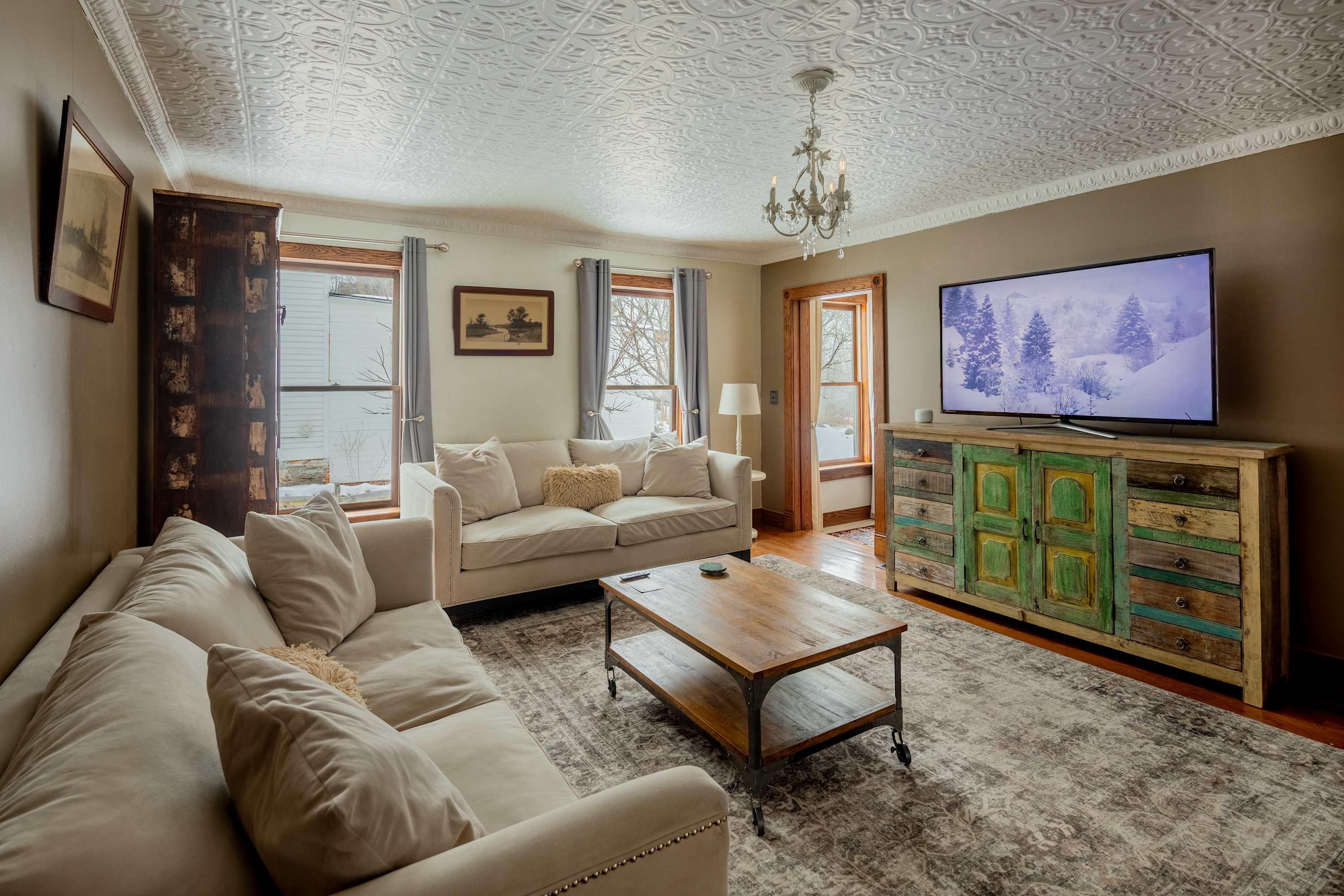 A bright living room features two cream-colored sofas, a rustic wooden coffee table, a large flat-screen TV on a colorful distressed cabinet, and a patterned area rug over hardwood floors.