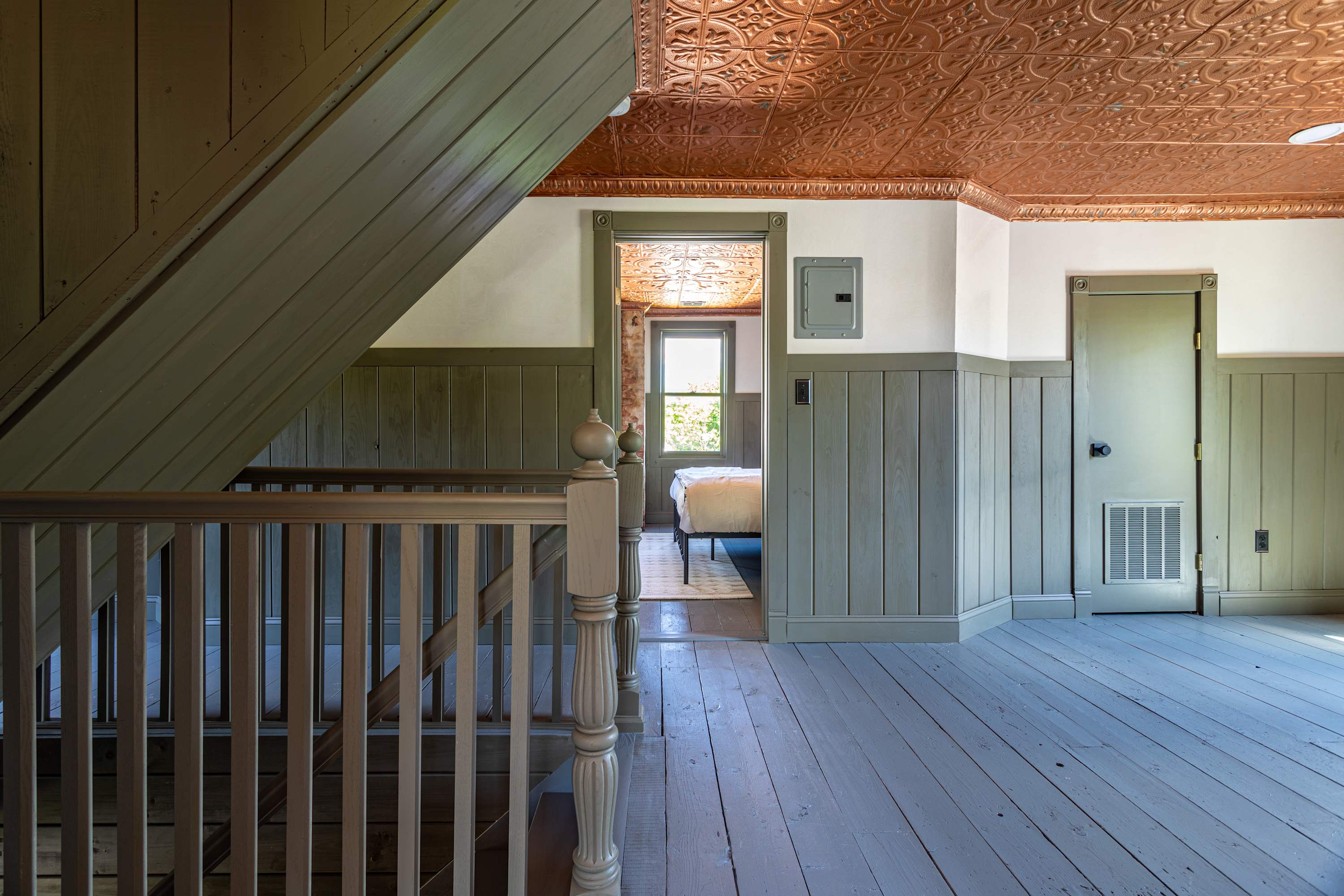 Upper floor landing with light green wood paneling, a wooden staircase railing, and a striking copper-colored pressed tin ceiling, with a glimpse into a bedroom.