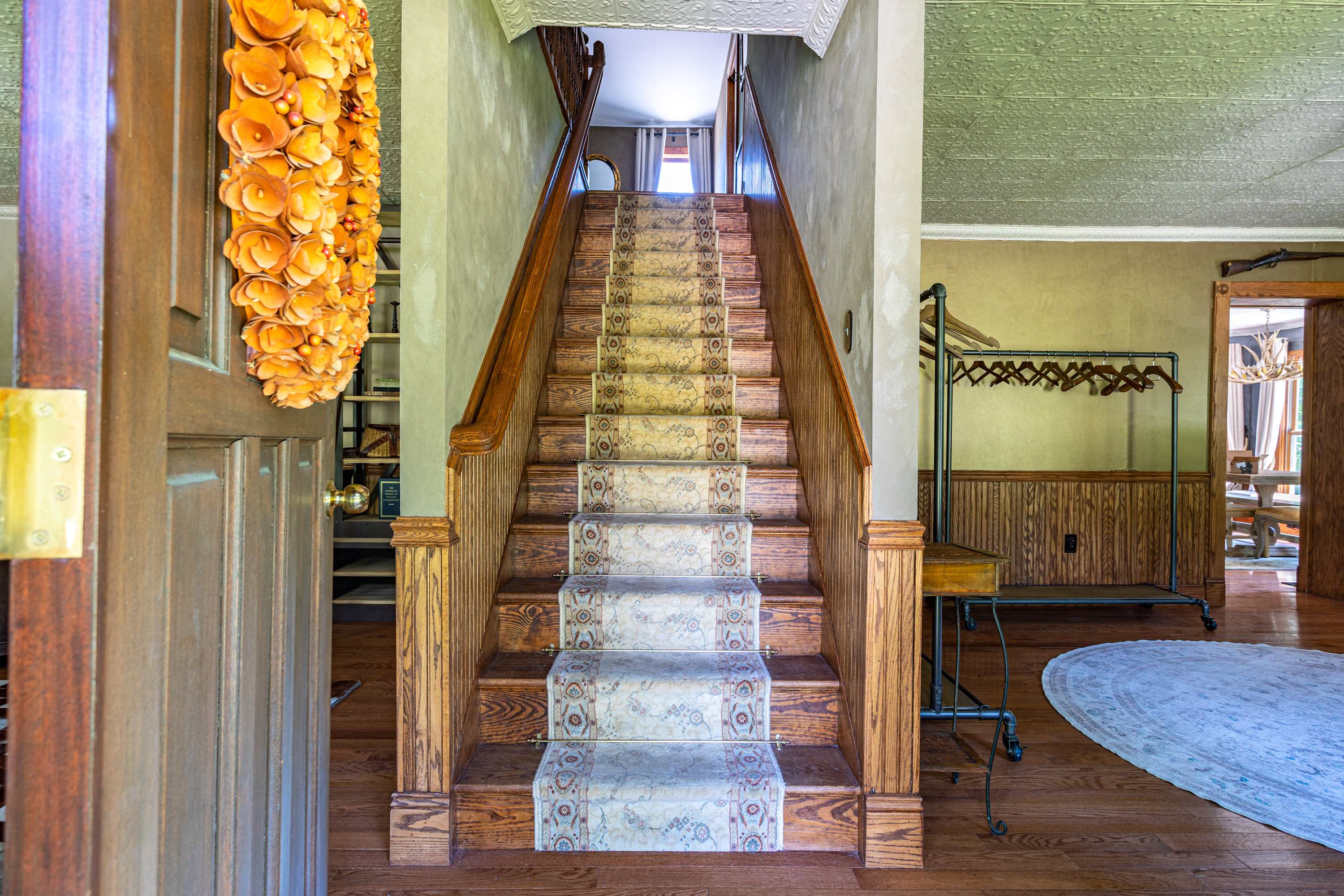 Grand foyer featuring a wooden staircase with a patterned runner, leading upstairs. The space has light green walls, wainscoting, and a decorative ceiling, with a coat rack visible to the right.