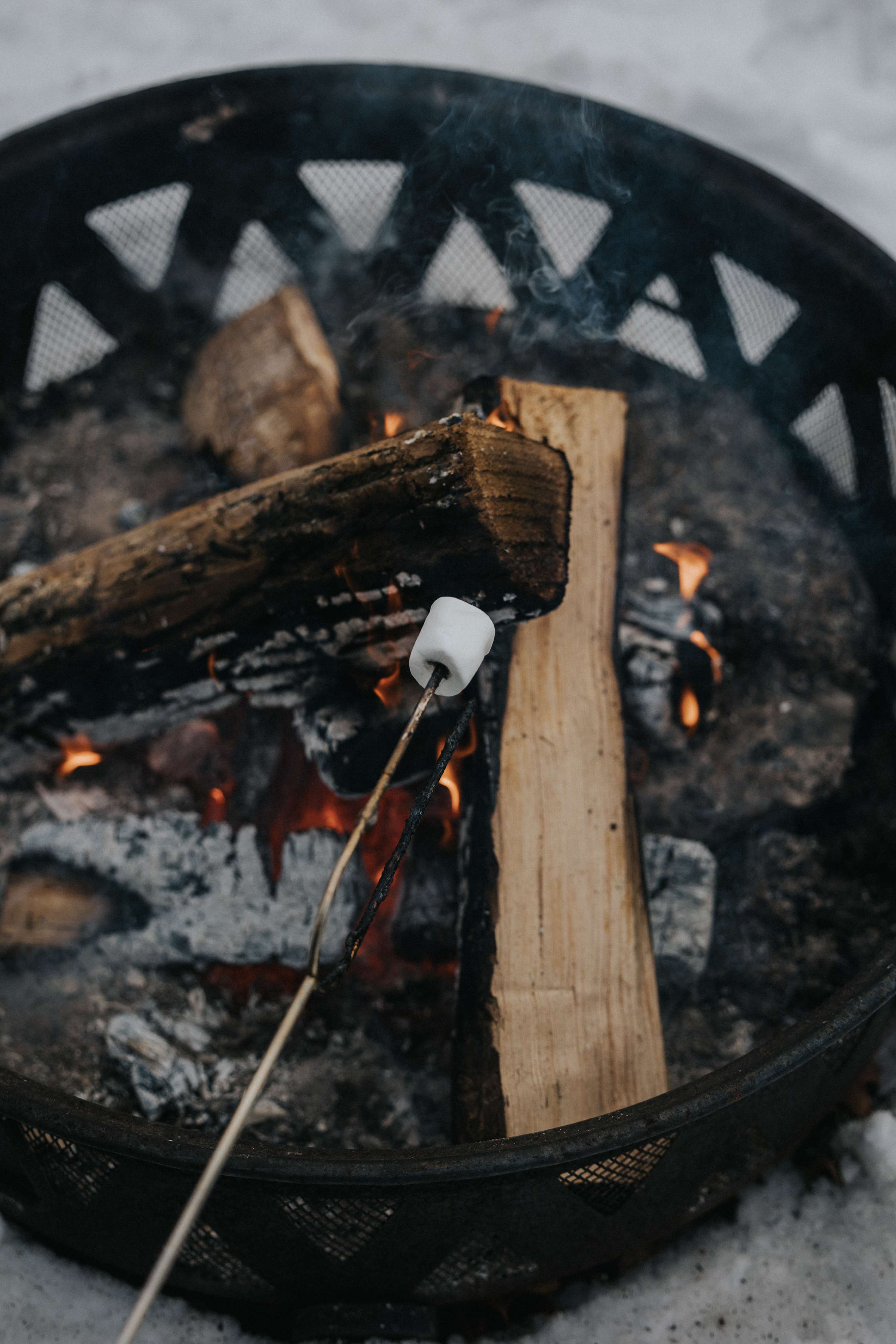 Close-up of a white marshmallow roasting on a stick over glowing embers and burning logs in a metal fire pit, with smoke gently rising.