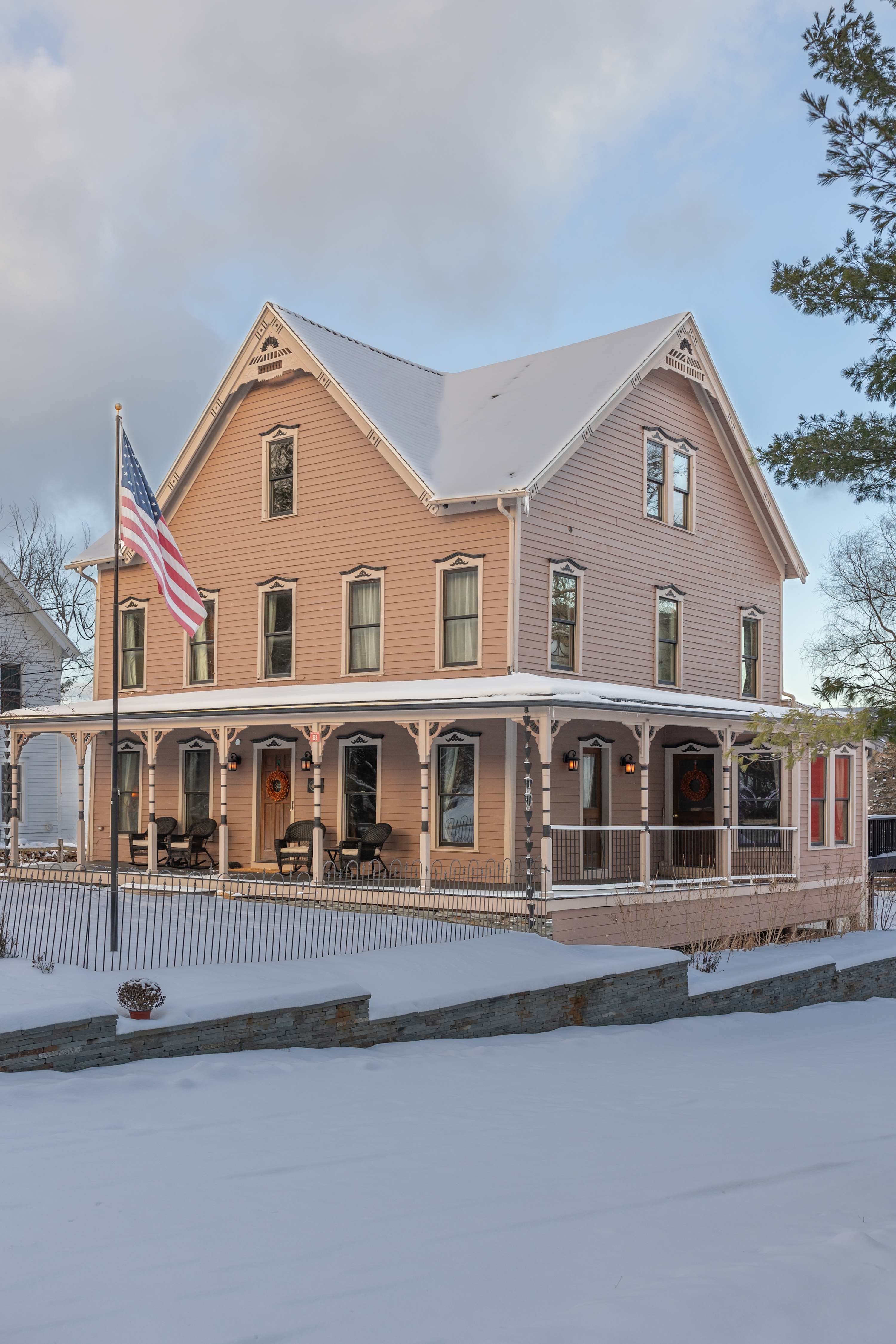 Charming two-story house with light pink siding and white trim, featuring a welcoming wrap-around porch adorned with wreaths and an American flag. The surrounding landscape is covered in a blanket of fresh winter snow.
