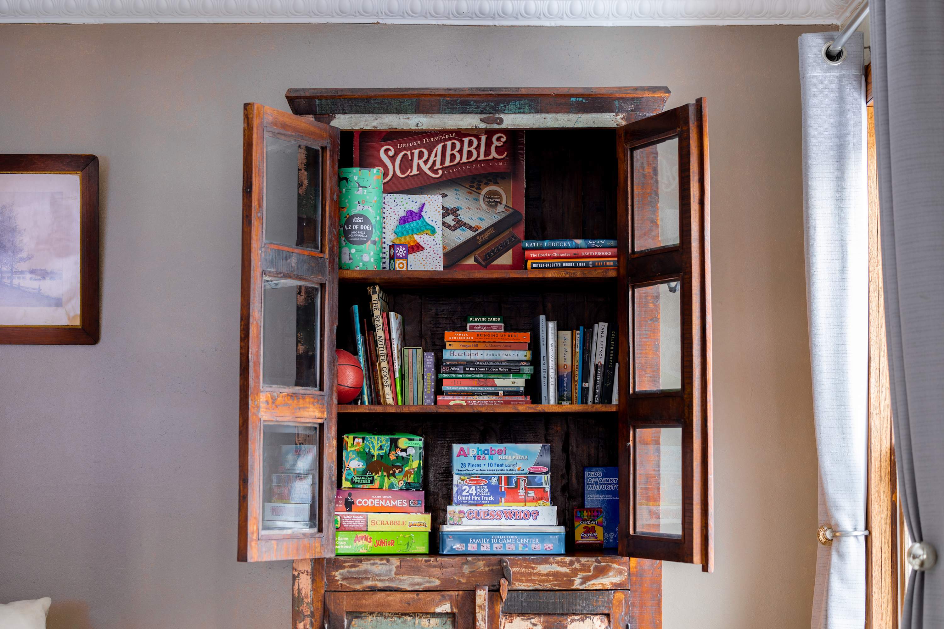Rustic wooden cabinet with glass doors, filled with a variety of board games, books, and small decorative items, suggesting a family-friendly entertainment space.