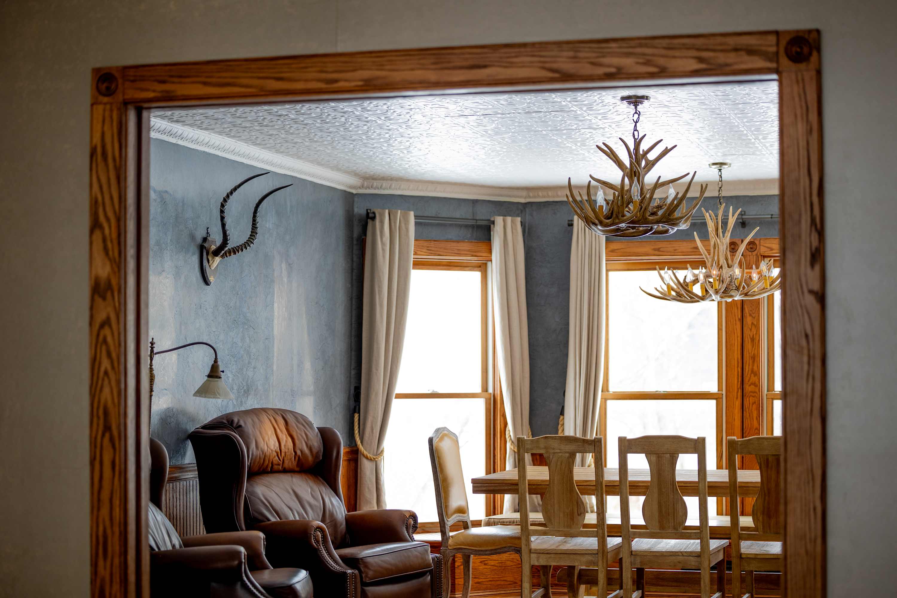 A view through a doorway reveals a dining room with a large wooden table and chairs, antler chandeliers, and a blue accent wall with a mounted animal skull, featuring large windows and a pressed tin ceiling.