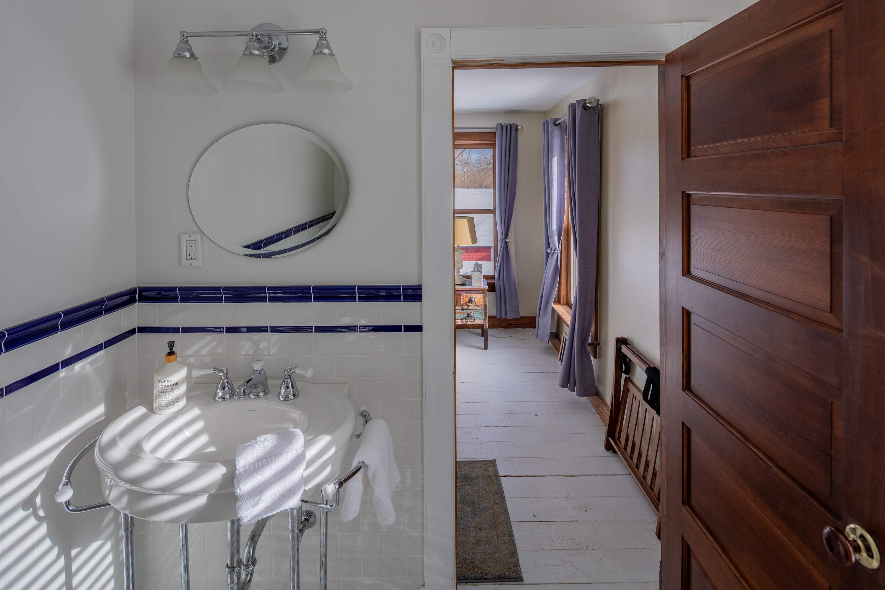 Bright bathroom featuring a pedestal sink with a round mirror, white subway tiles with blue accent stripes, and a view into an adjacent bedroom through an open doorway.