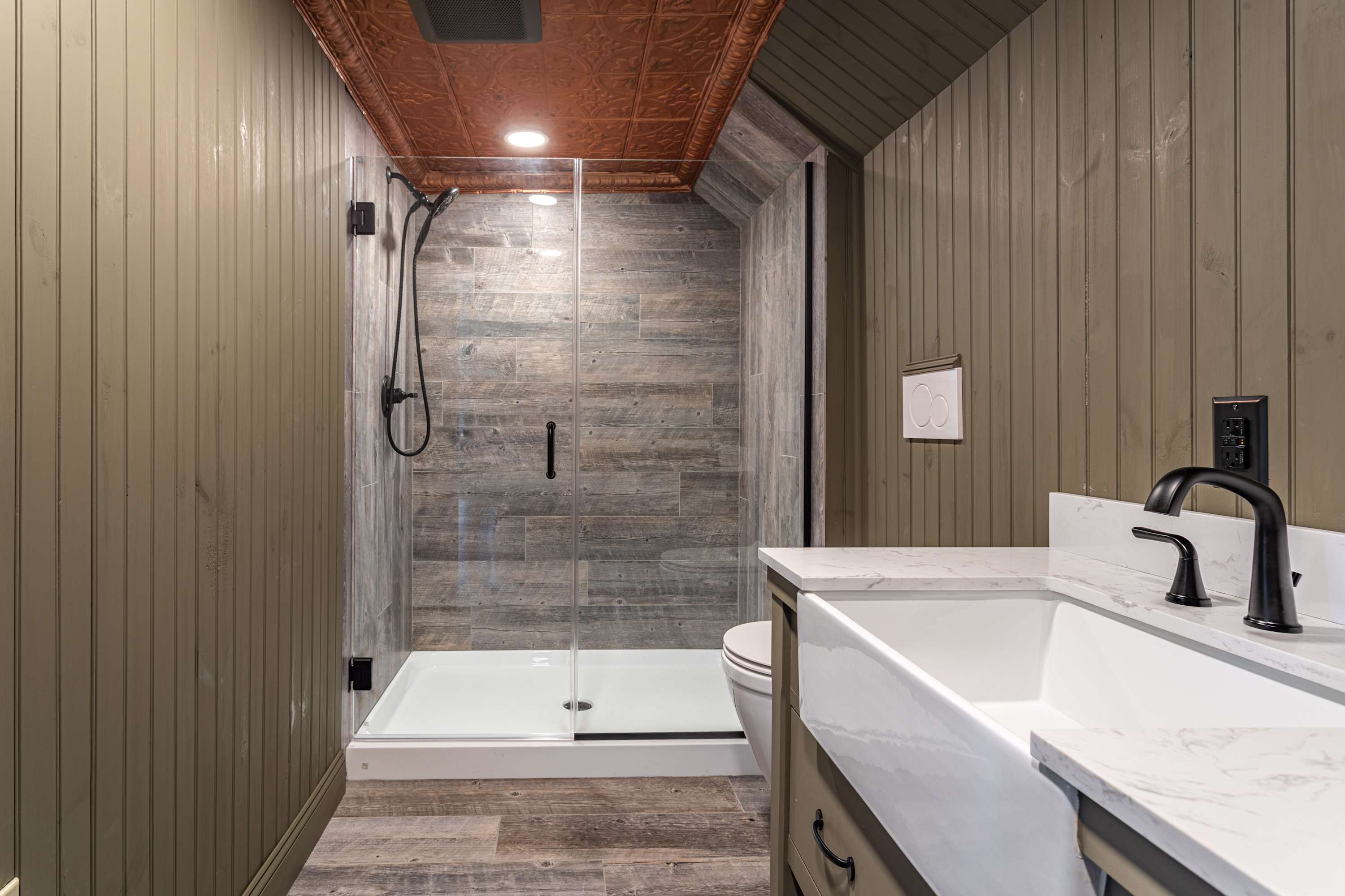 Spacious bathroom with a walk-in shower featuring wood-look tile, a modern toilet, and a double vanity with white farmhouse sinks and marble countertops, surrounded by wood paneling.