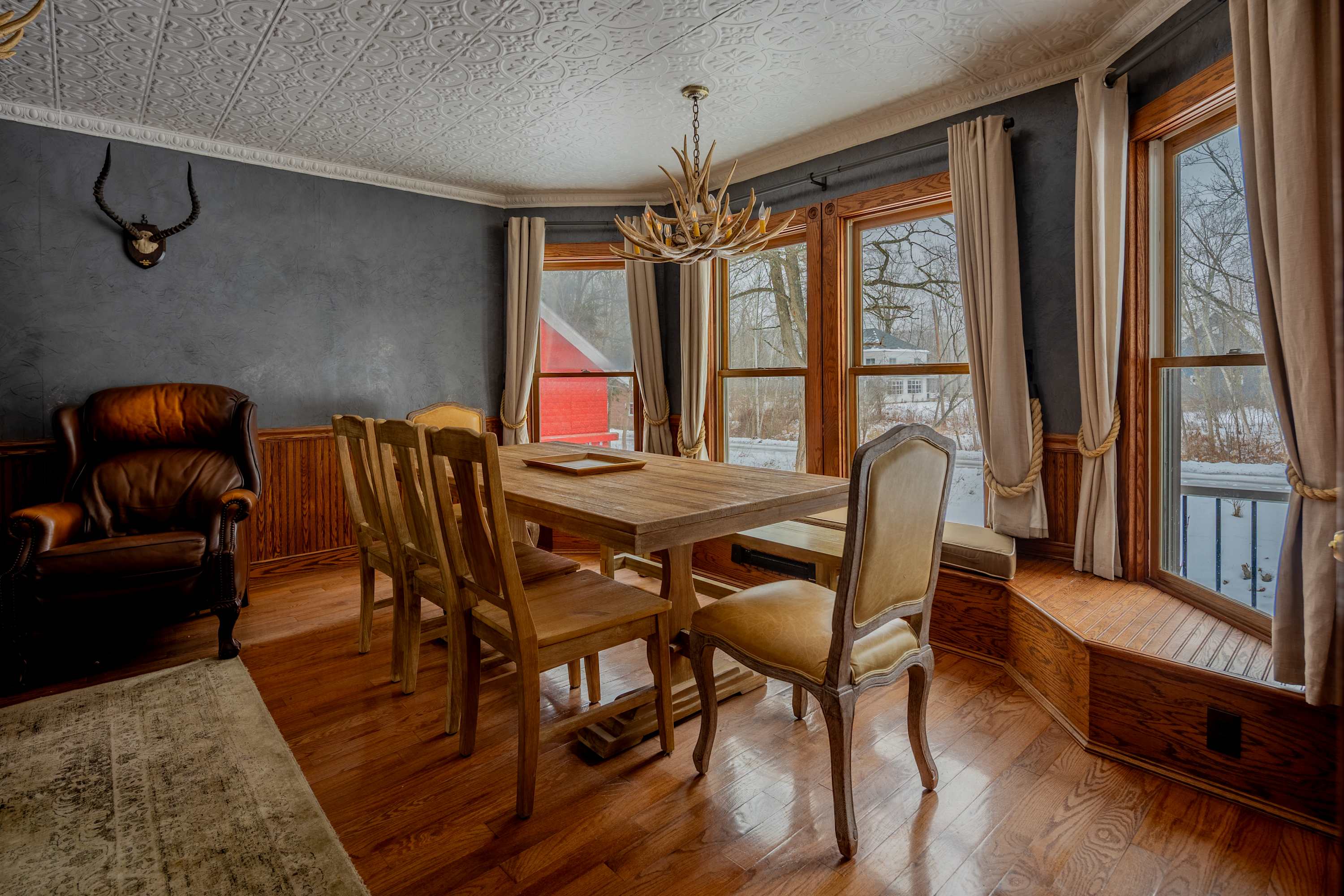 A spacious dining room with a long wooden table and matching chairs, a brown leather armchair, and a blue accent wall with a mounted animal skull, featuring large windows overlooking a snowy landscape.
