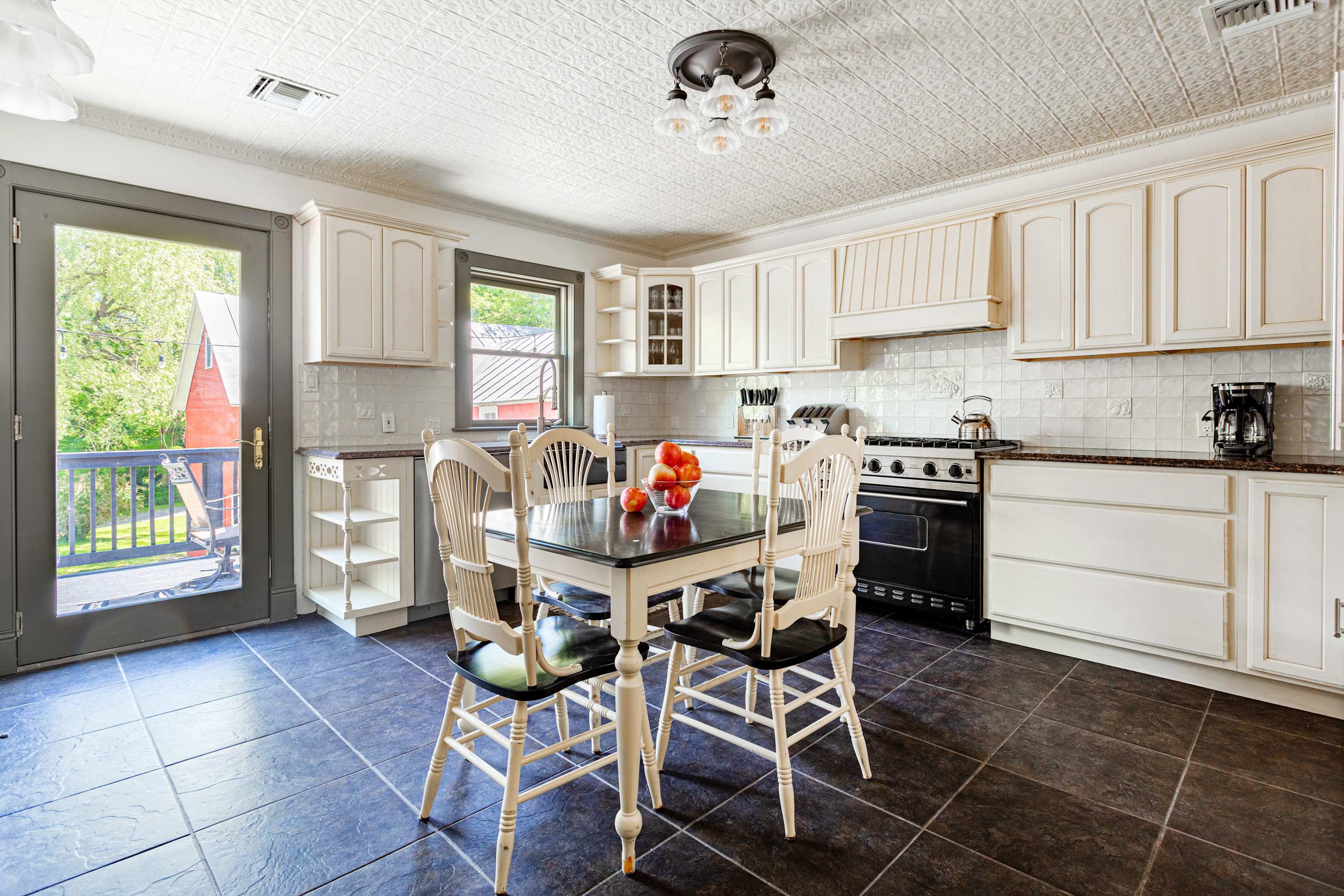 Bright kitchen with white shaker cabinets, dark countertops, and a black range. A black dining table with white chairs sits on dark tiled floors, with a glass door leading to an outdoor area.