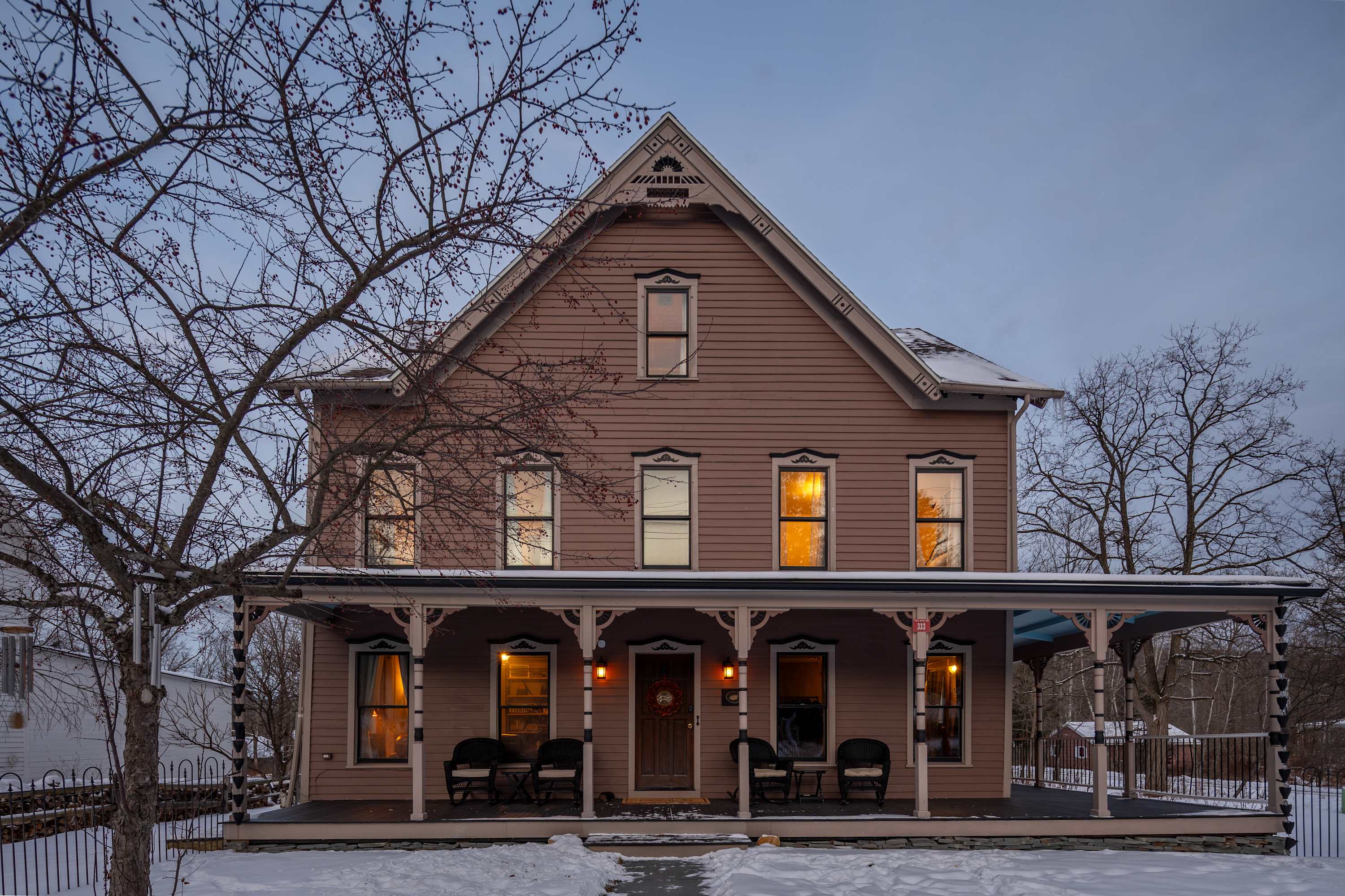 Front exterior of a large, traditional house with light brown siding and a wide porch, illuminated by warm interior lights at dusk. Snow covers the ground and bare trees frame the scene.
