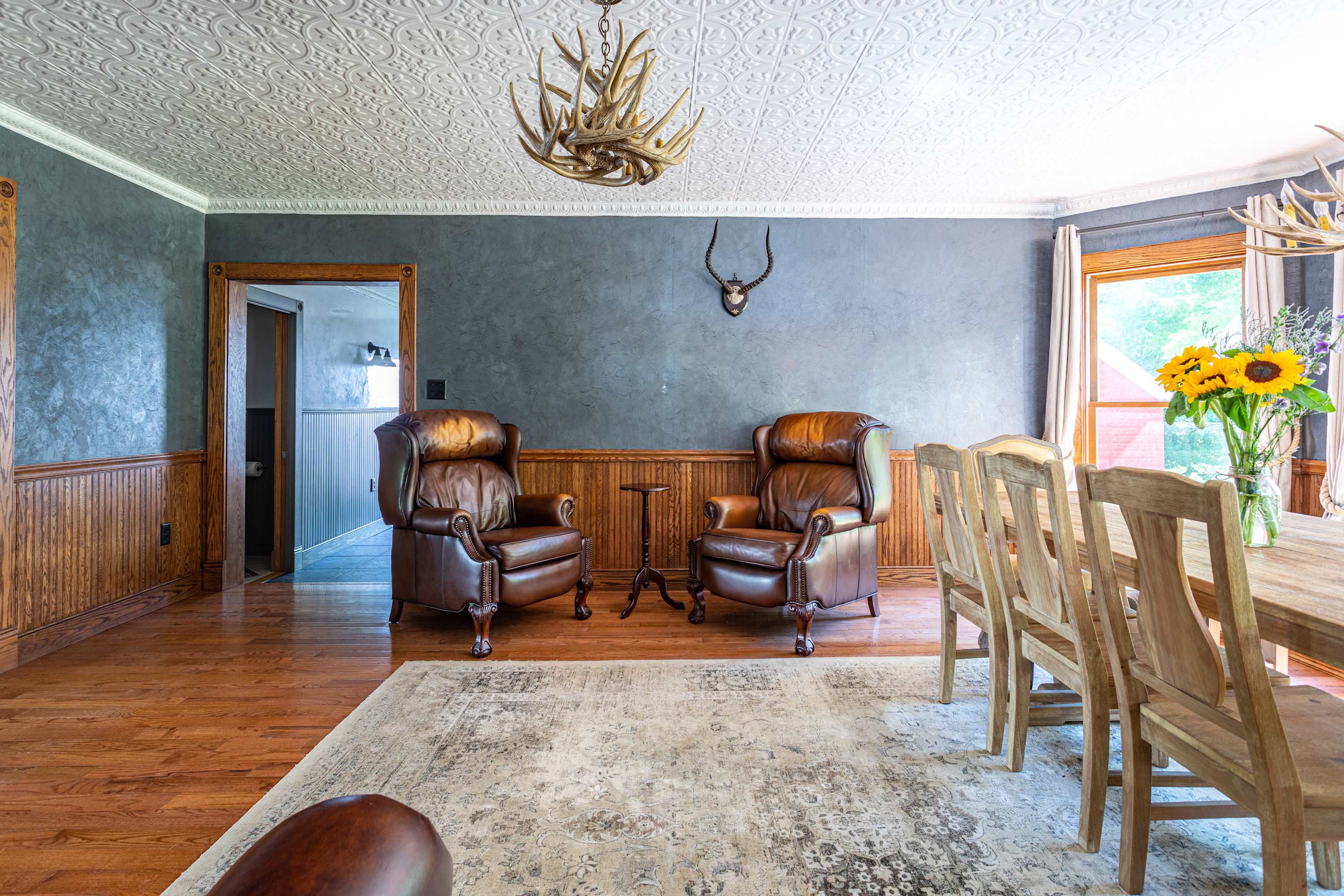 Rustic dining room with a large wooden dining table and chairs, complemented by two brown leather armchairs. The room features dark gray walls, wooden wainscoting, hardwood floors, and an antler chandelier.