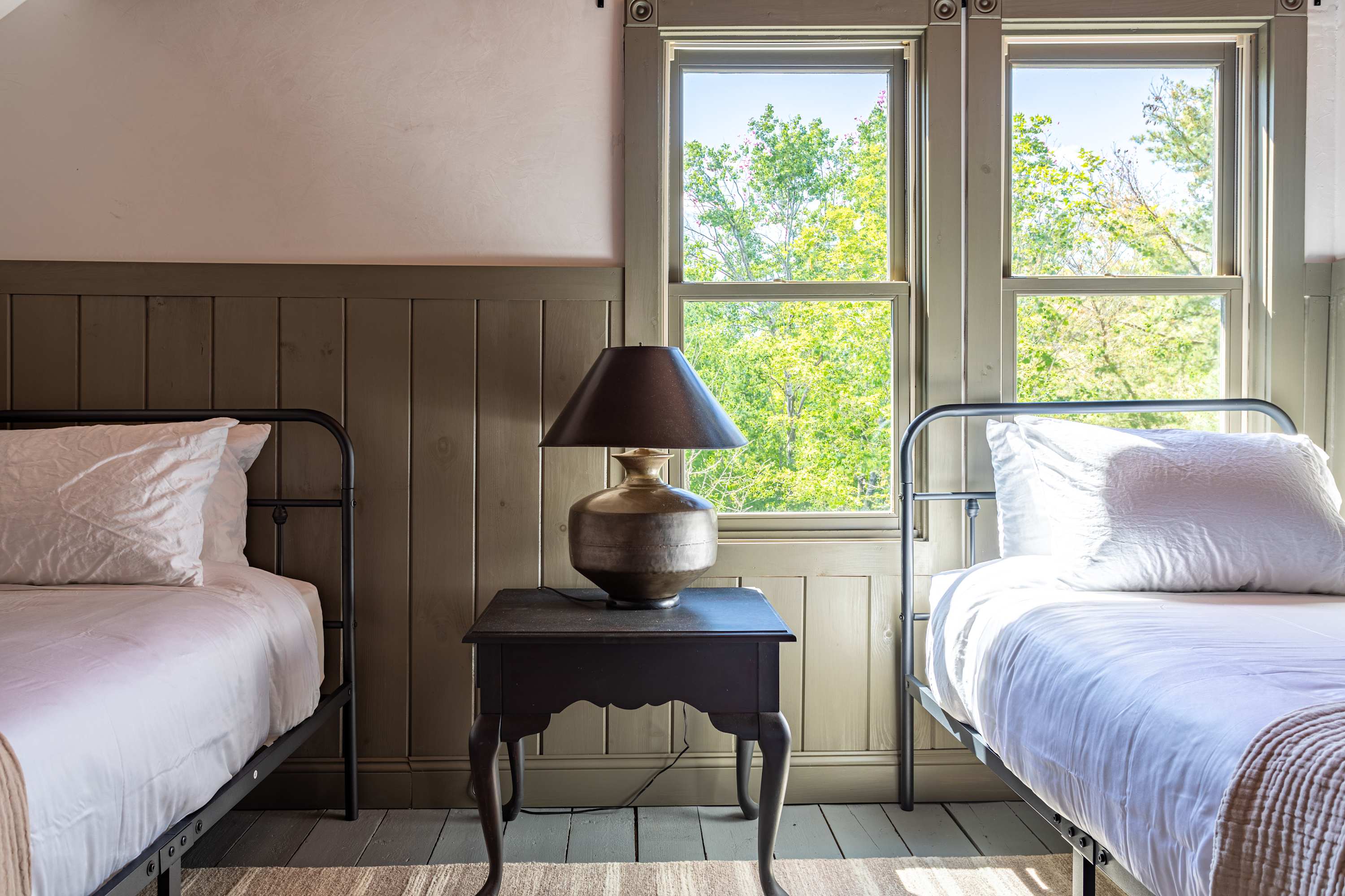 Close-up view of two black metal frame twin beds separated by a dark wooden nightstand with a bronze table lamp, set against light green wood paneling and large windows.