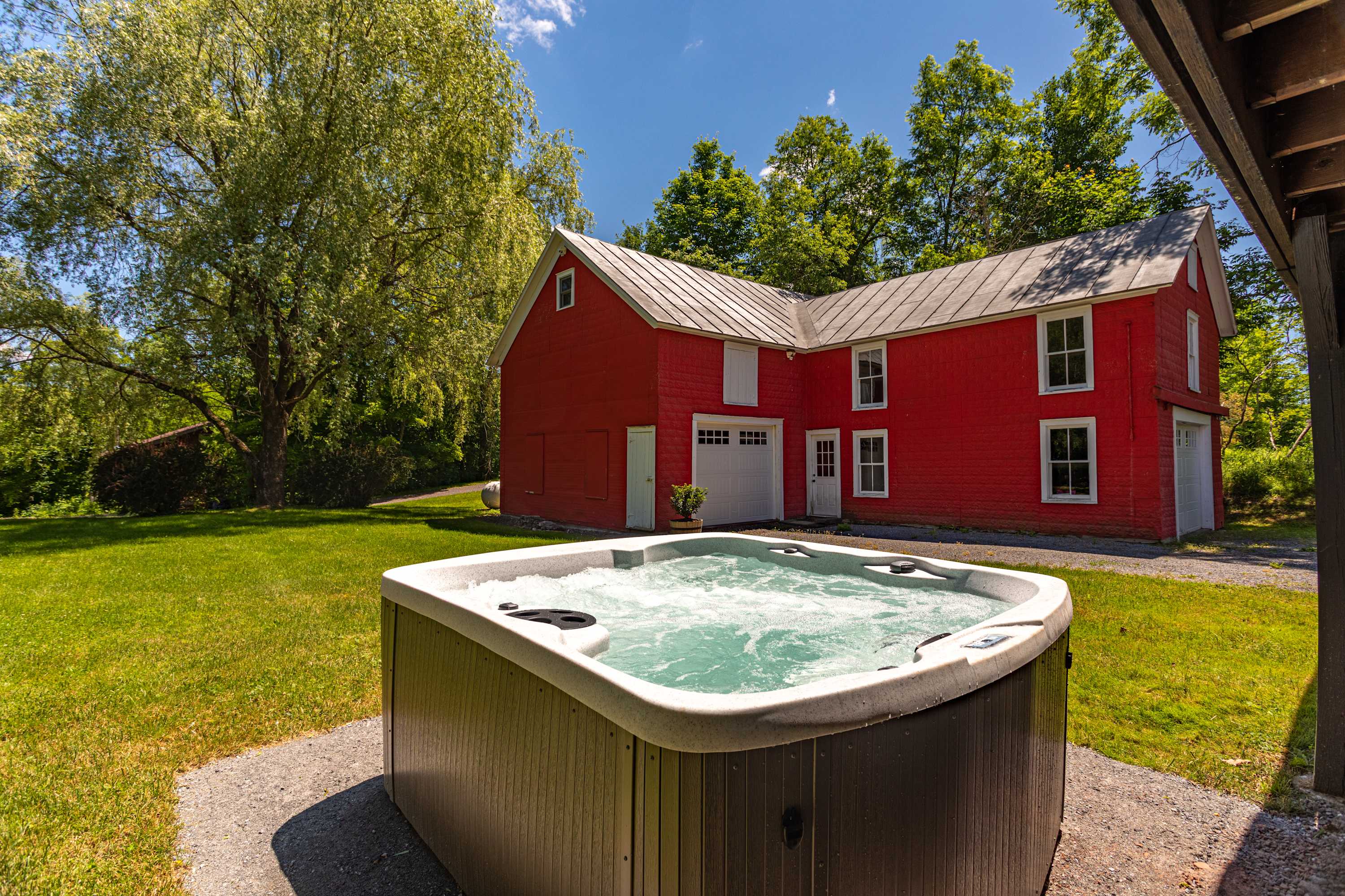 Outdoor hot tub with bubbling water, set on a gravel patio amidst a lush green lawn. In the background, a vibrant red barn with white trim and multiple windows stands under a clear blue sky.