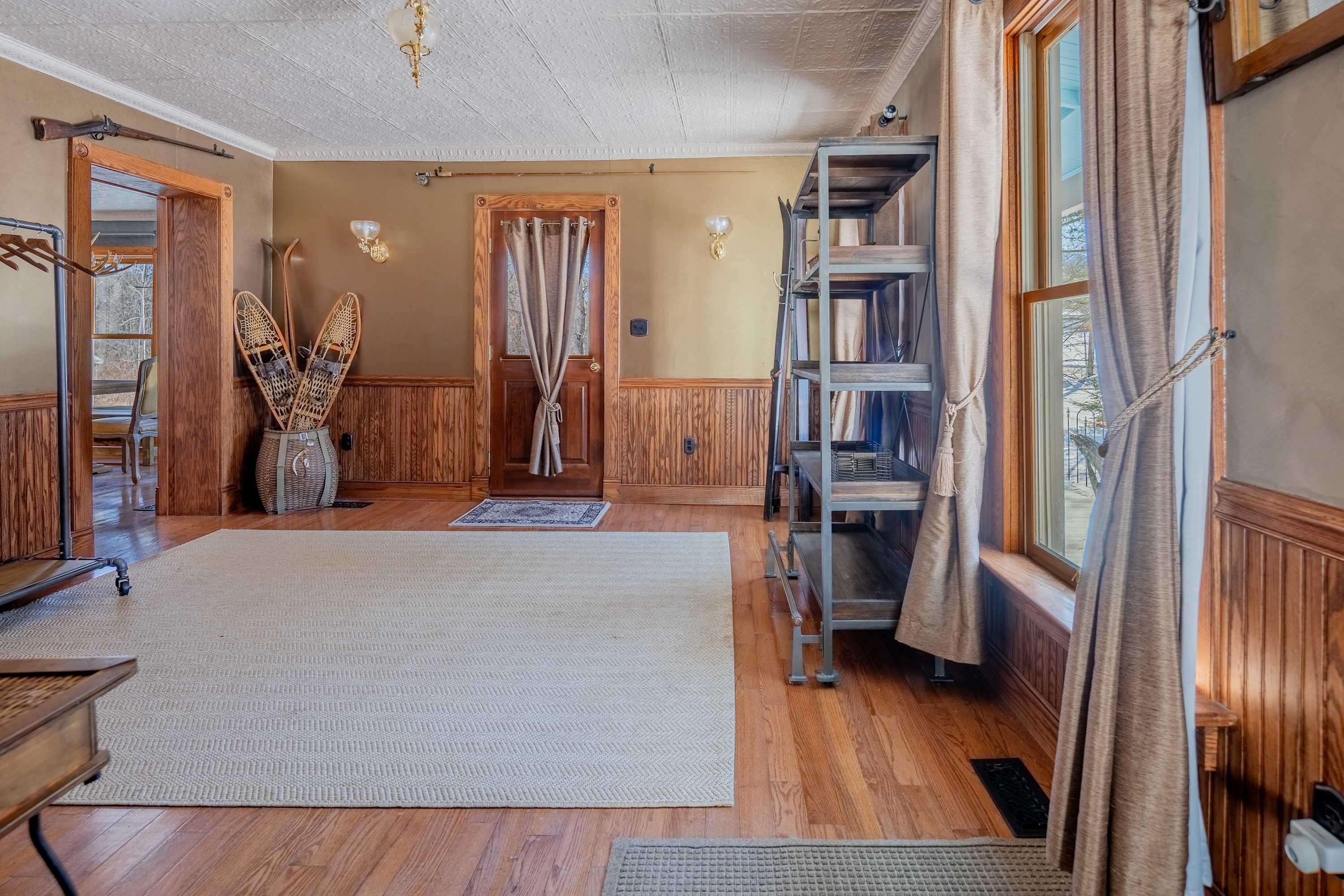 Warm and inviting foyer featuring polished hardwood floors, dark wood wainscoting, and a decorative white ceiling. A tall metal shelving unit stands beside a large window, and a pair of snowshoes decorates a corner.