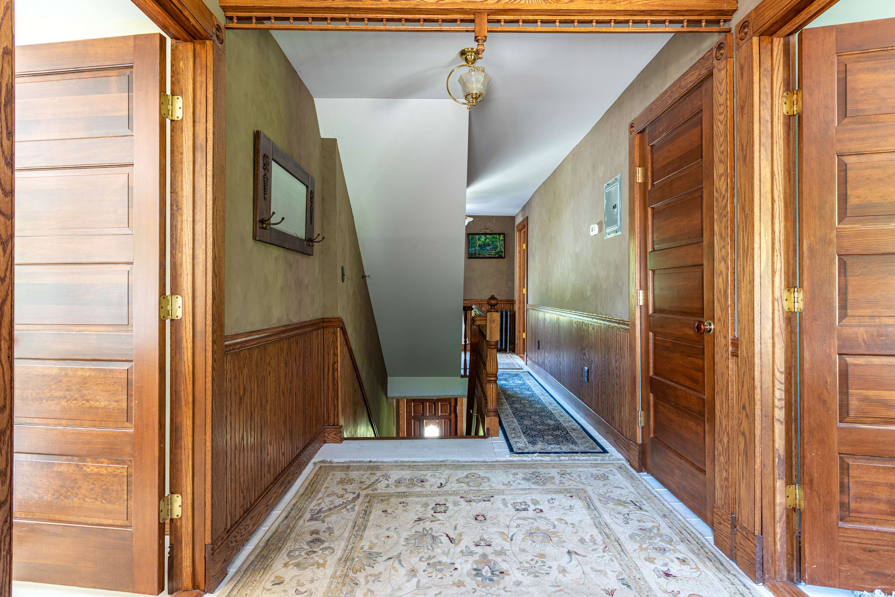 Grand foyer with rich wooden double doors, wainscoting, and a patterned area rug leading to a staircase and a long hallway with another rug.