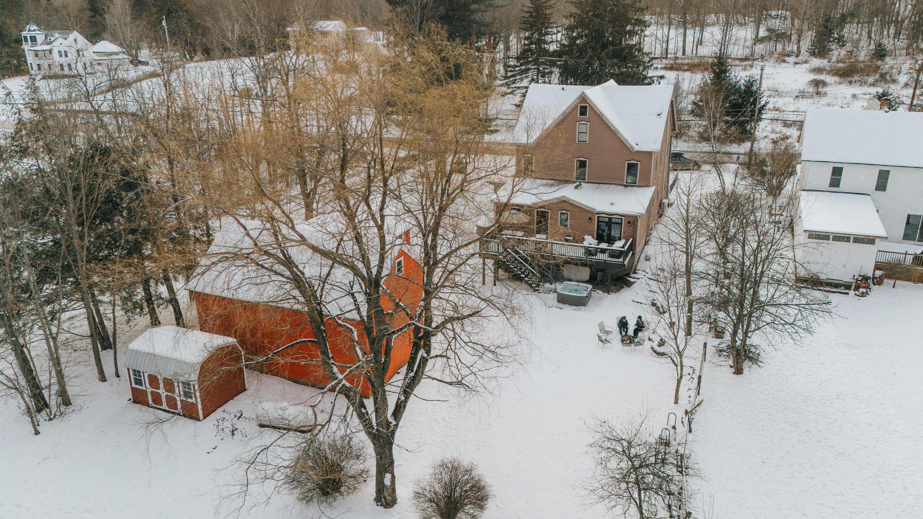 An aerial view of the property in winter, showing the main house with a deck and hot tub, a red barn, and a fire pit area, all surrounded by snow-covered trees and a rural setting.