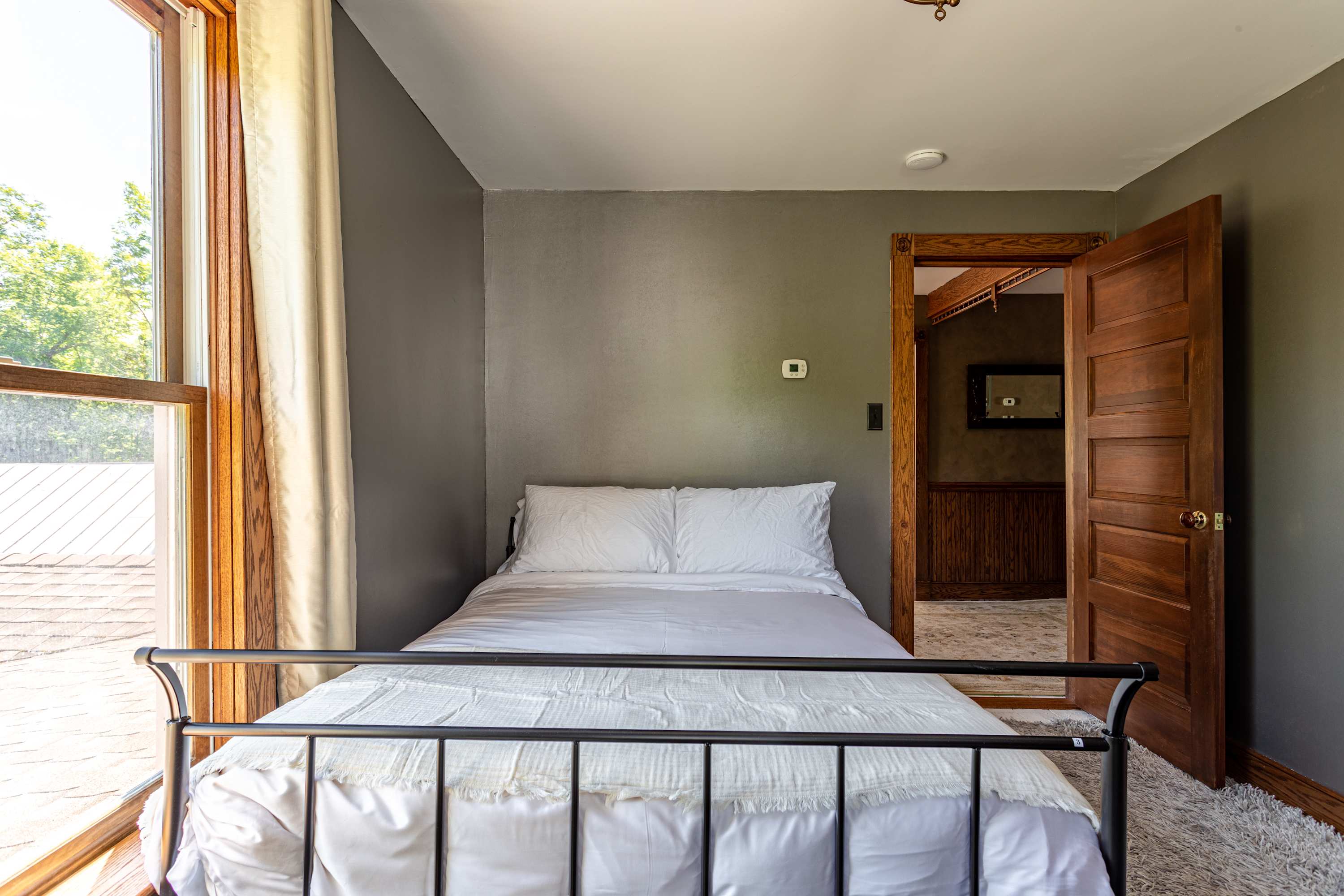 Cozy bedroom featuring a black metal frame queen bed with crisp white bedding, dark gray walls, and a large window with light curtains offering views of the outdoors.