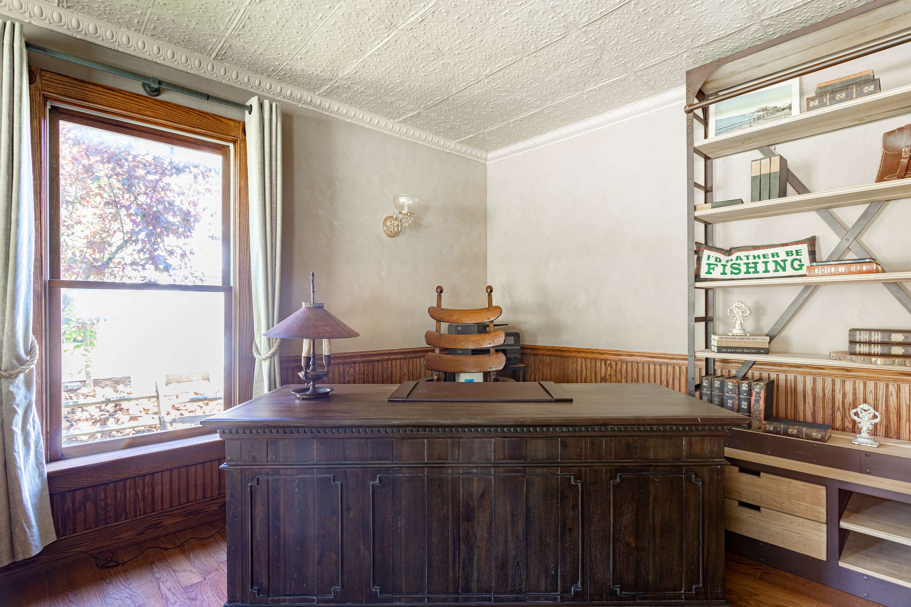 Detailed view of a classic wooden desk with a vintage lamp and chair, positioned by a large window with wooden trim, in an office with wood paneling and a white pressed tin ceiling.