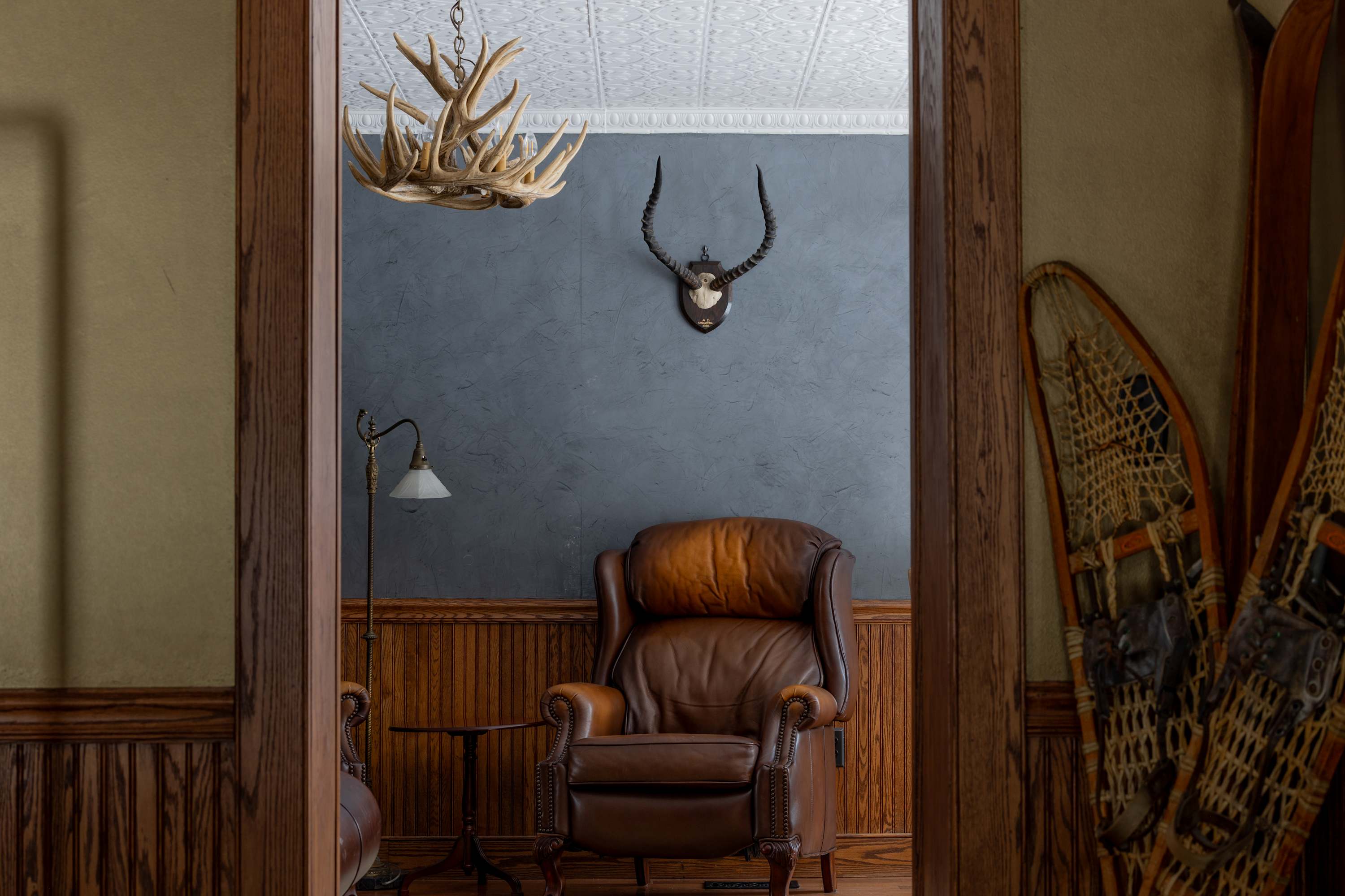 A cozy sitting area features a large brown leather armchair, a small side table with a lamp, and a wall adorned with a mounted animal skull, all framed by dark wood trim and a pressed tin ceiling.