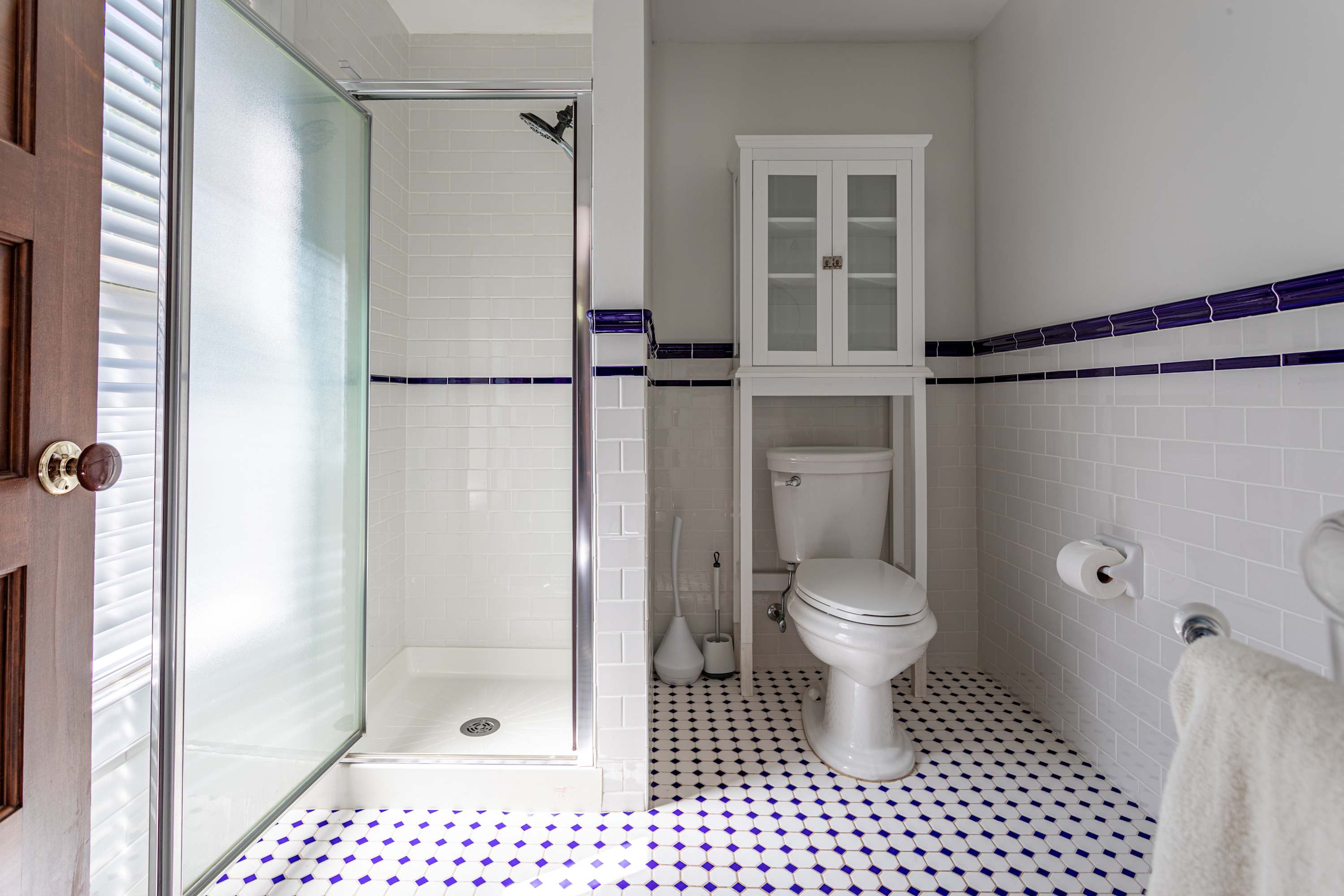 Modern bathroom with white subway tiles, a glass-enclosed shower, toilet, and a white cabinet above the toilet, all set against a blue and white patterned floor.