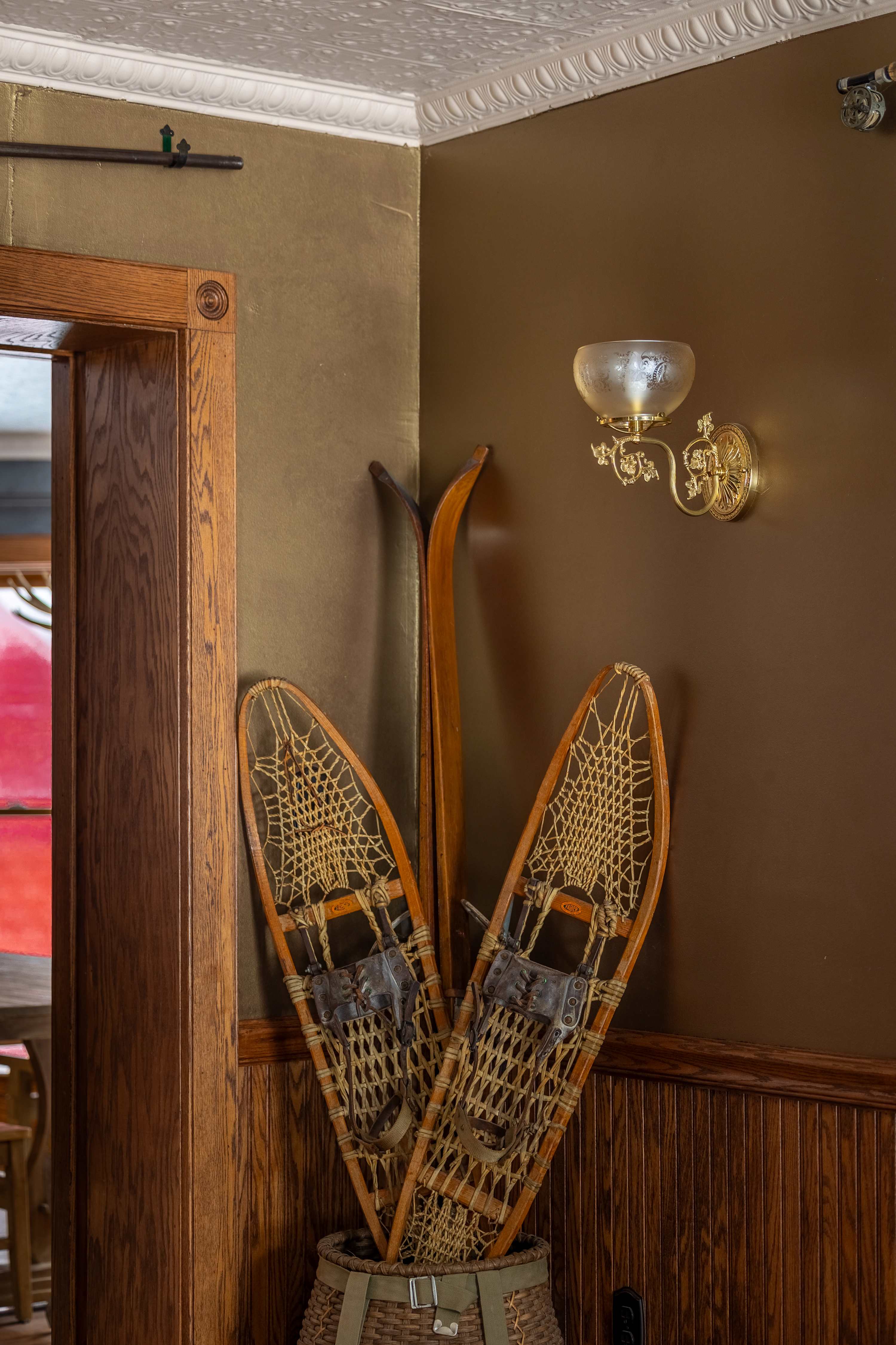 A corner of a room features vintage wooden snowshoes and skis leaning against a wall with dark wood wainscoting and a warm brown paint color, illuminated by an ornate gold wall sconce.