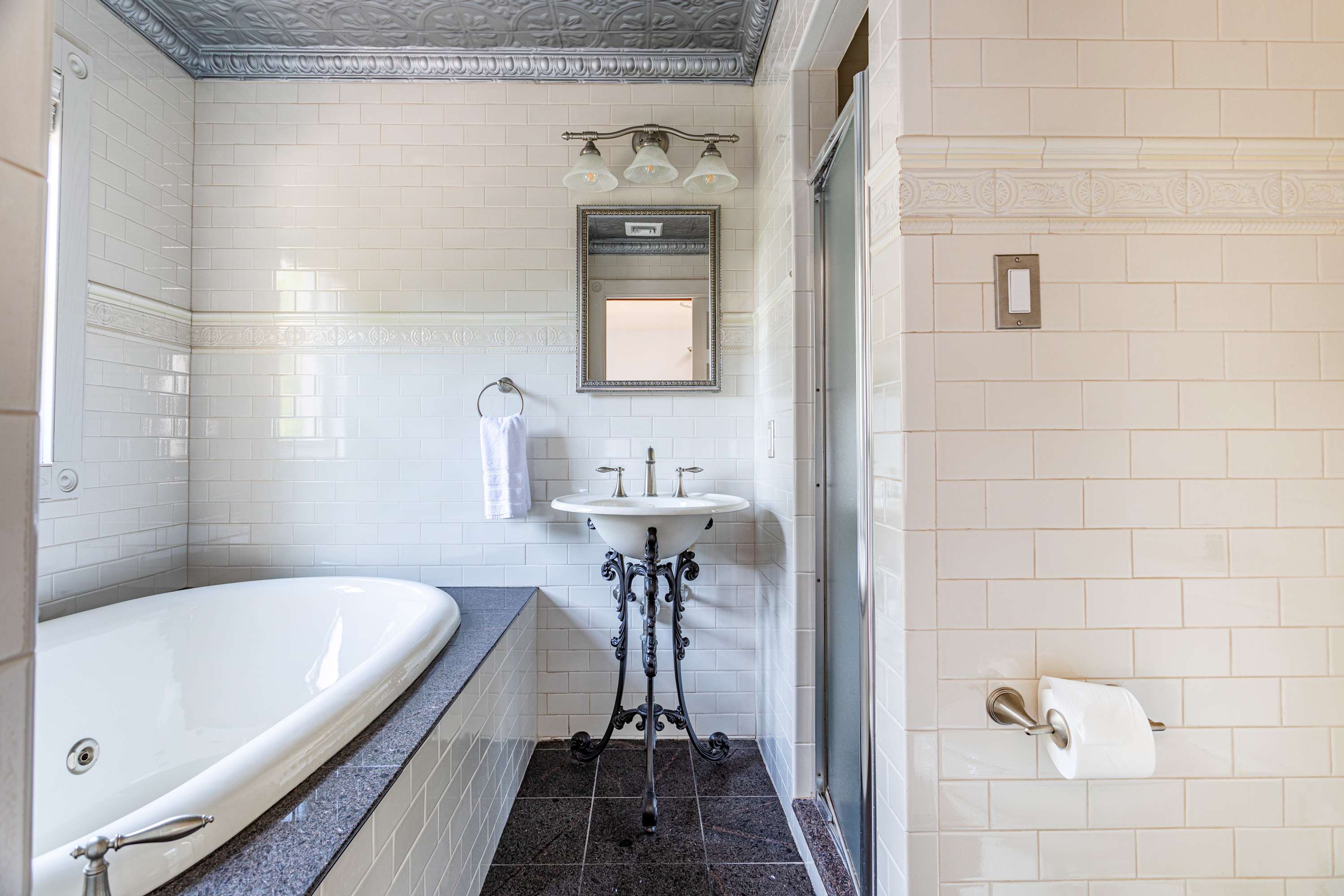 Elegant bathroom with white subway tile walls, a jetted bathtub with dark granite surround, and a pedestal sink. A glass-enclosed shower is visible, along with a mirrored medicine cabinet.