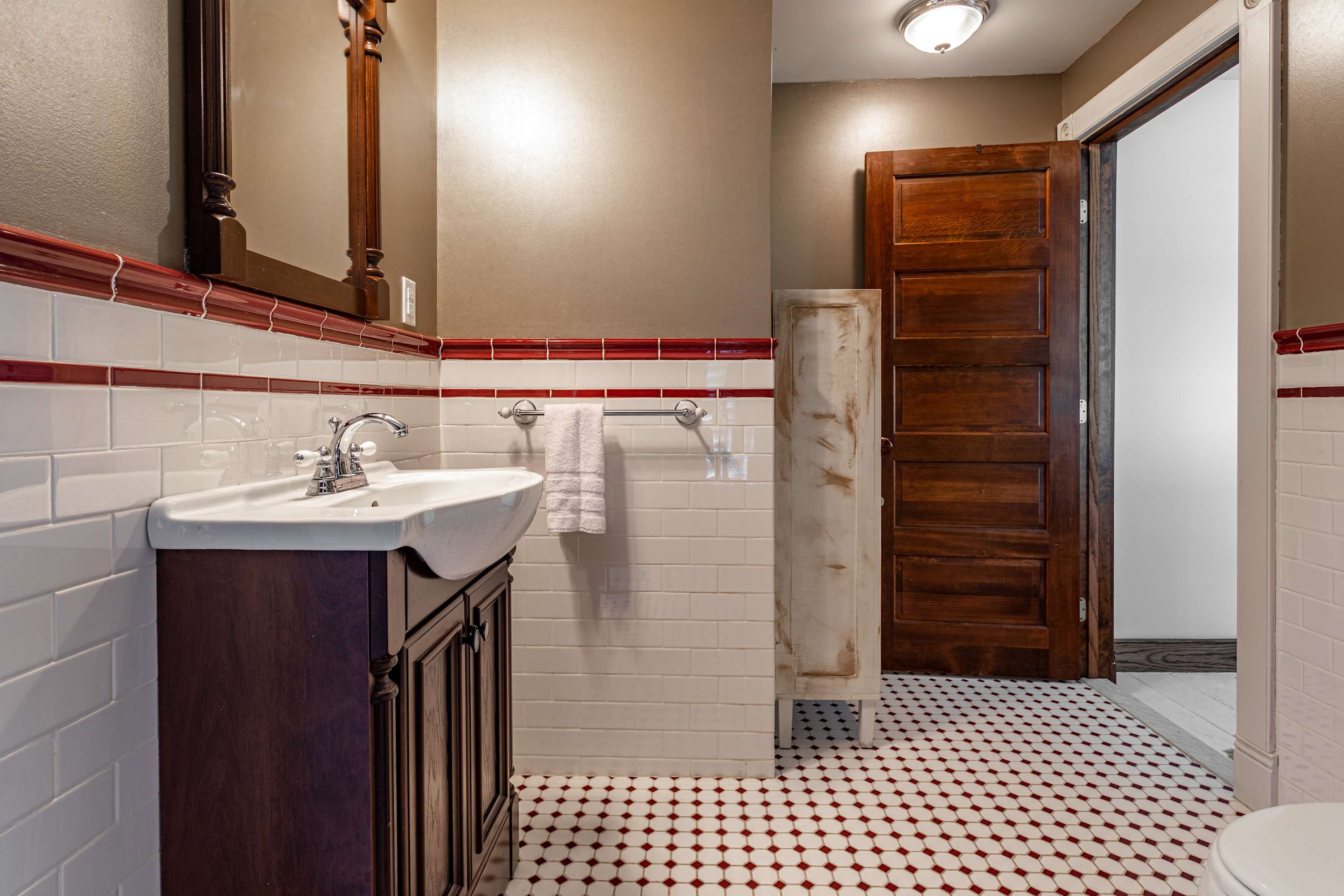 Bathroom with a dark wood vanity and sink, white subway tiles with a red accent stripe, and red and white patterned floor tiles, with a wooden door visible.