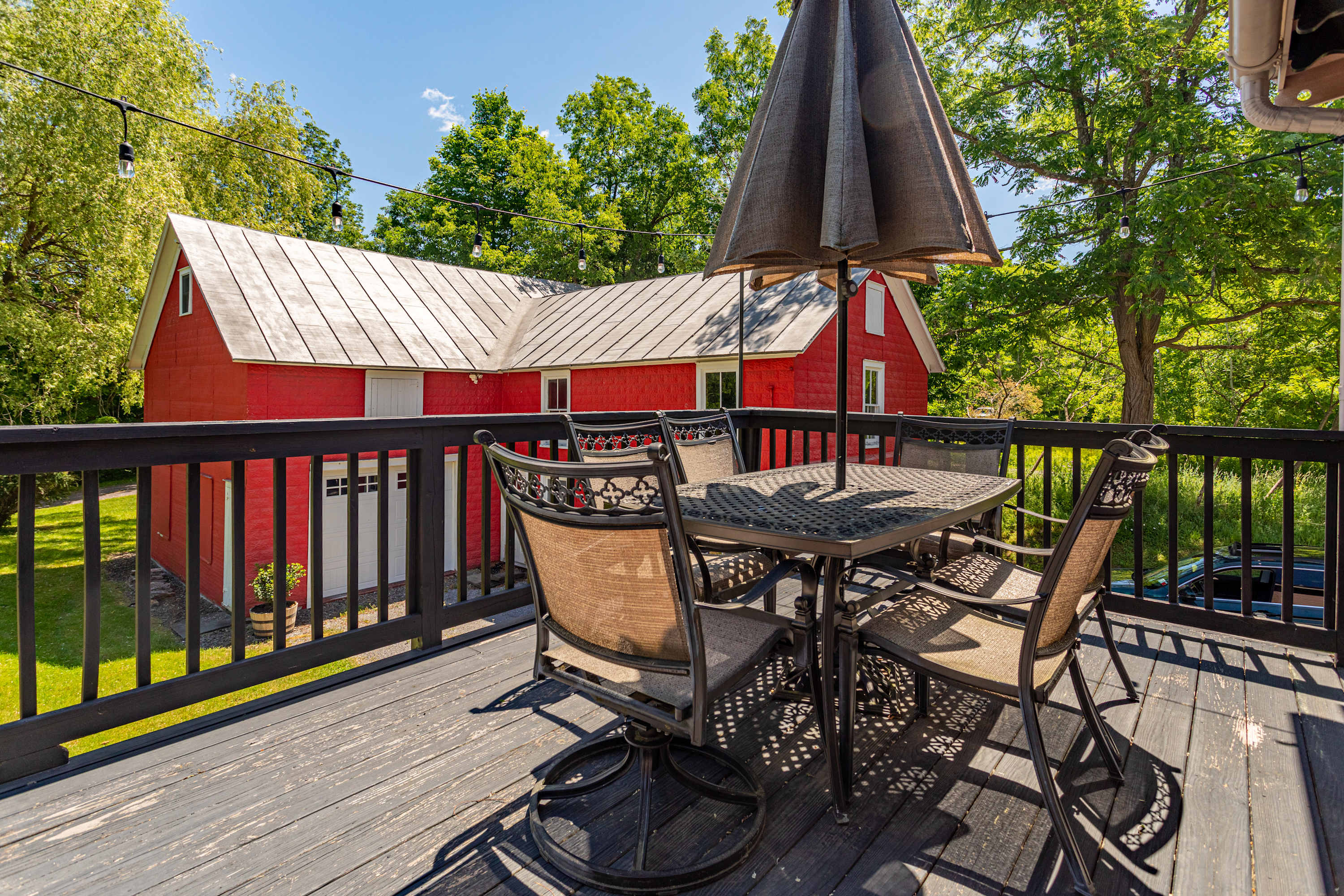 Outdoor deck with a dark metal dining table and chairs, shaded by an umbrella, offering views of a vibrant red barn with a silver roof and surrounding green trees under a clear sky.