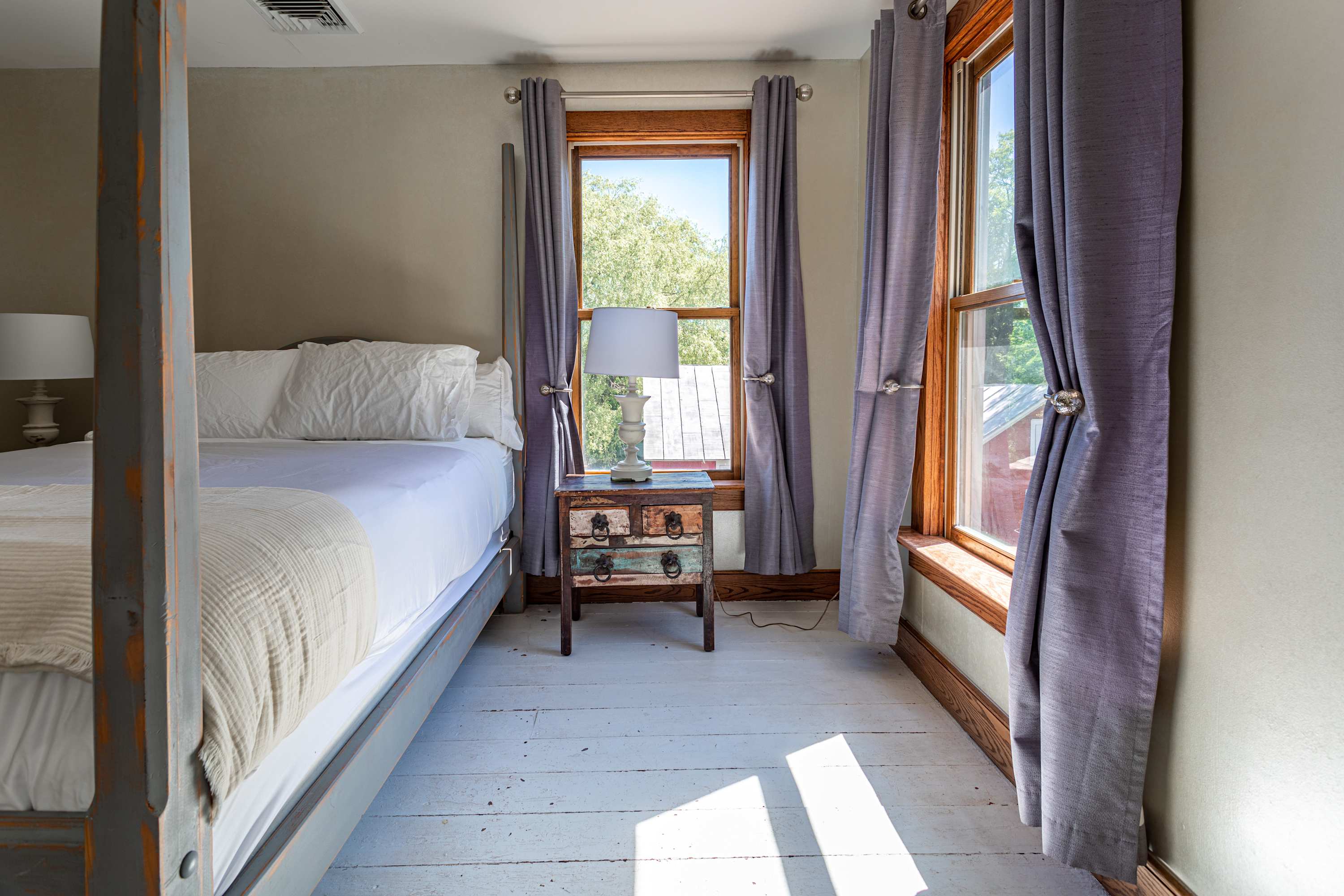 Bright bedroom featuring a four-poster bed with white bedding, distressed white wood floors, and two large windows with grey curtains offering views of trees.