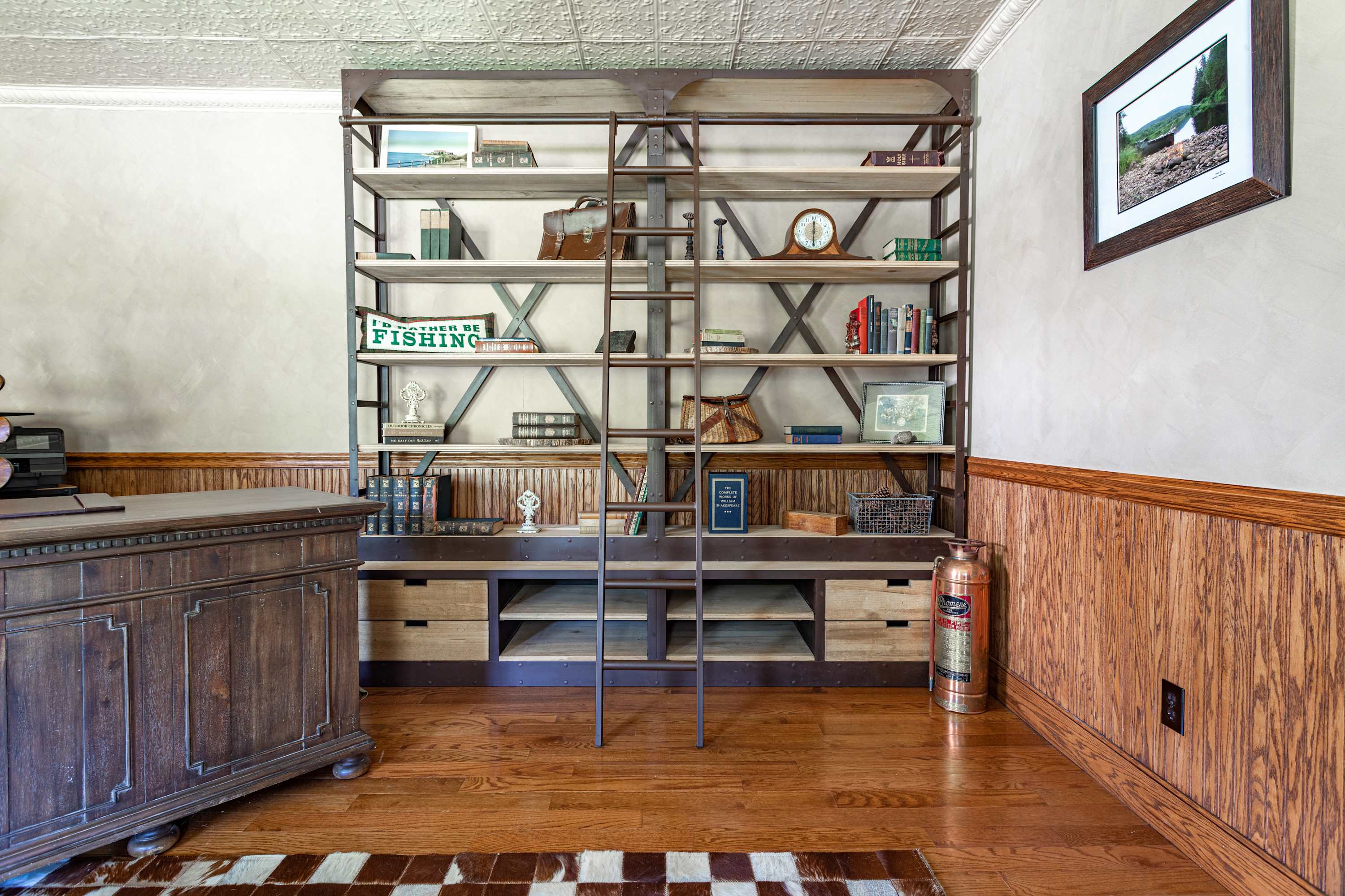 Elegant office space featuring a large industrial-style bookshelf with a ladder, a substantial wooden desk, and rich wood paneling on the lower walls, topped with a white pressed tin ceiling.