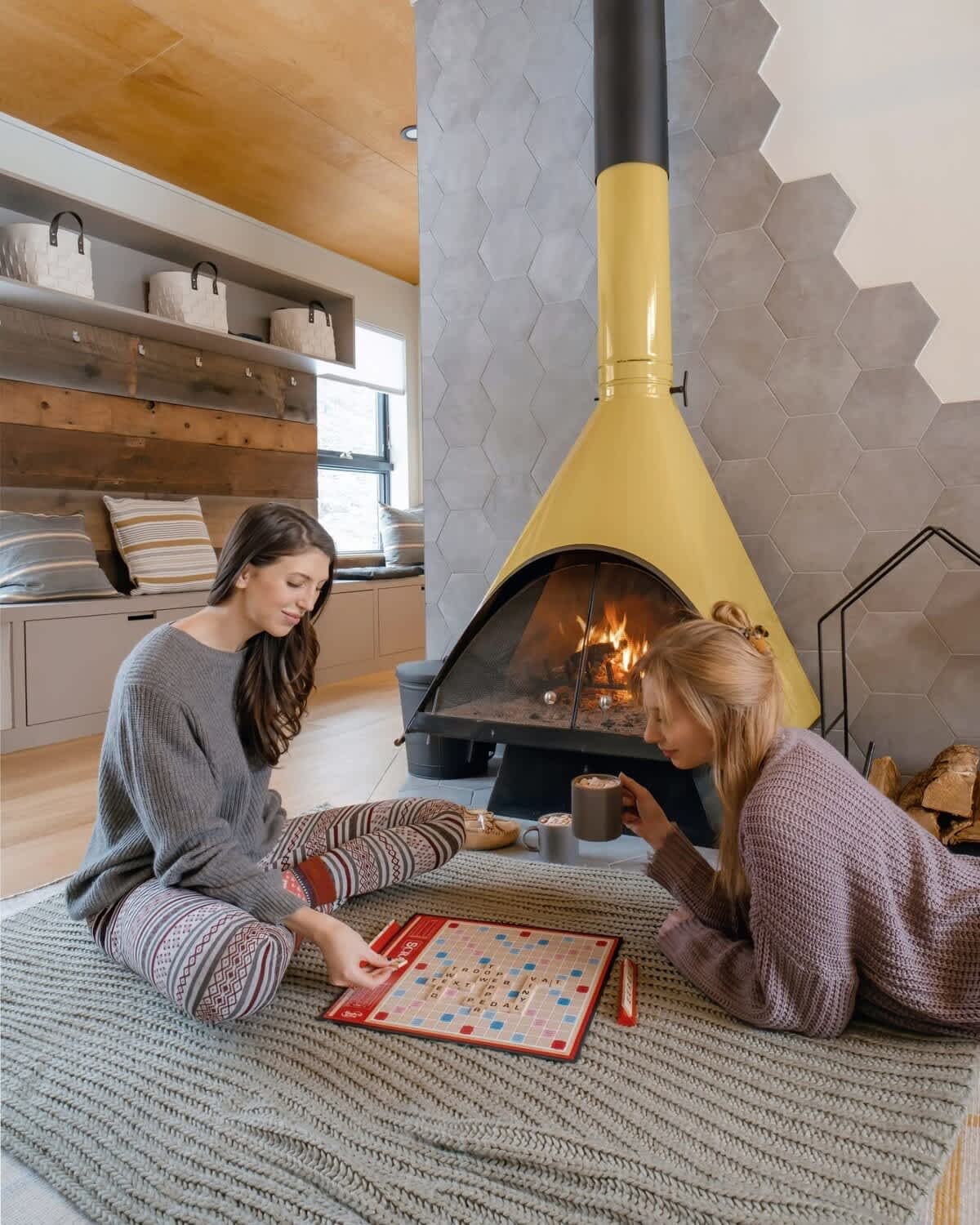 Two women are comfortably seated on a textured rug, playing Scrabble in front of a lit yellow freestanding fireplace. The cozy living space features a warm wood-paneled ceiling and a built-in window seat with storage and decorative baskets.