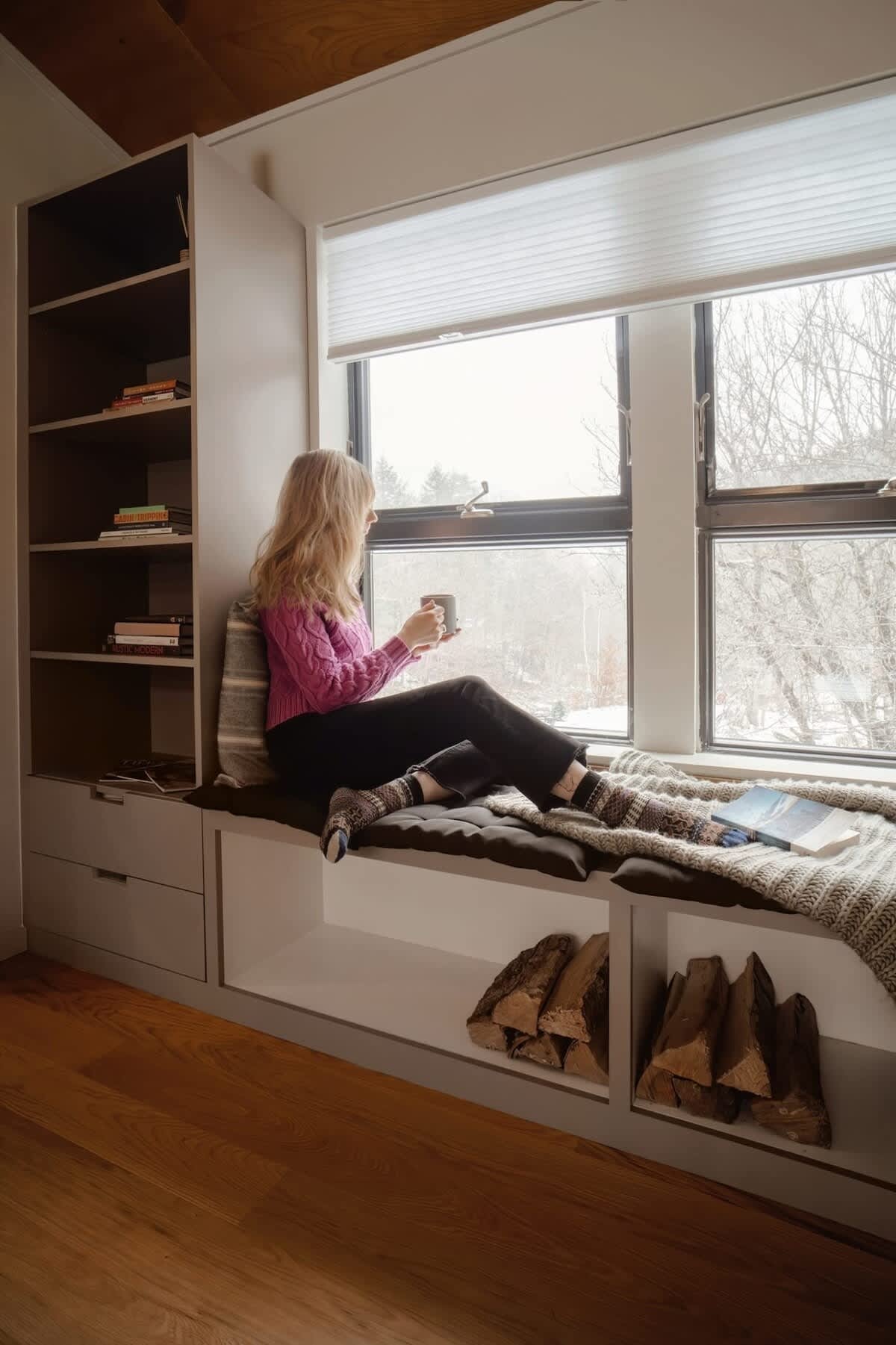 A cozy window seat area with built-in gray shelving, where a person in a pink sweater and dark pants sits comfortably, looking out the window with a mug. Stacked firewood is visible in the storage compartments below the seat.