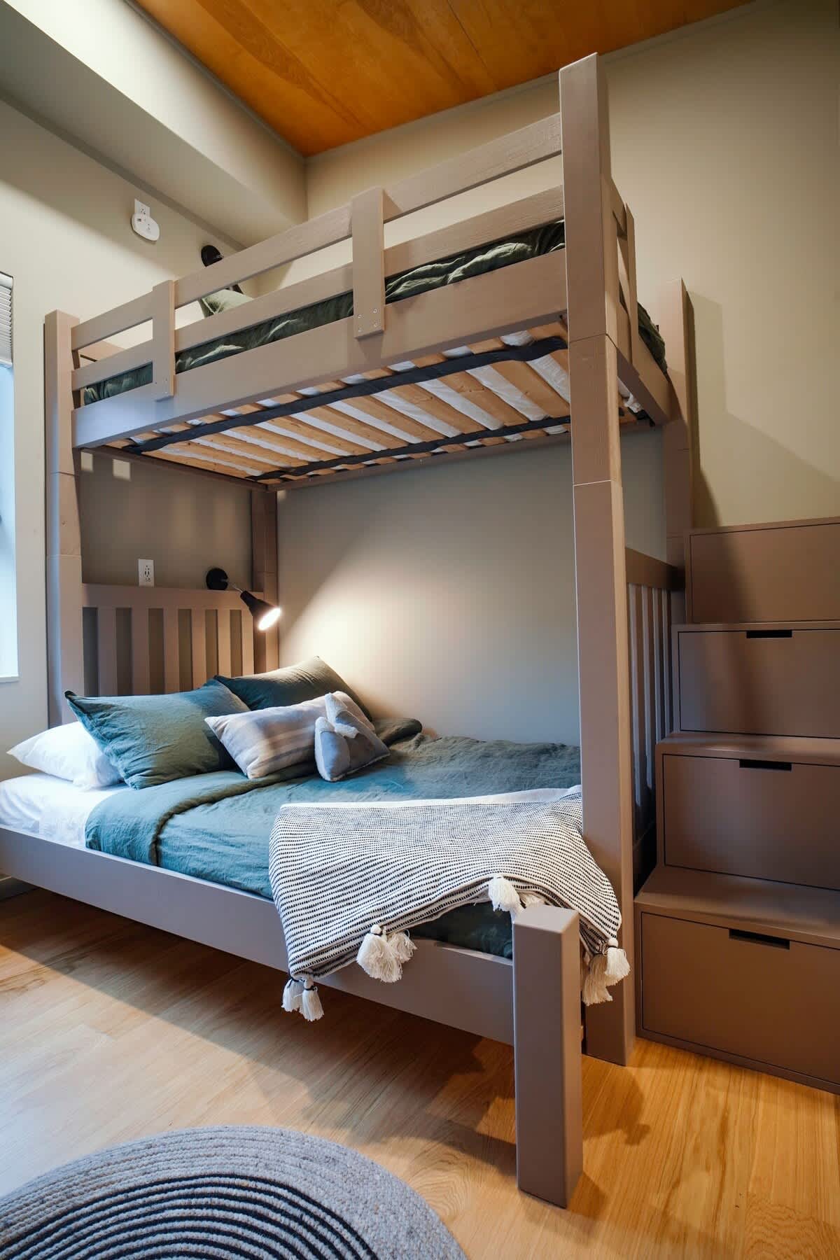 Close-up view of the wooden bunk bed in bedroom-2, showing the green bedding, decorative pillows, and the integrated storage steps. A round rug is on the floor.