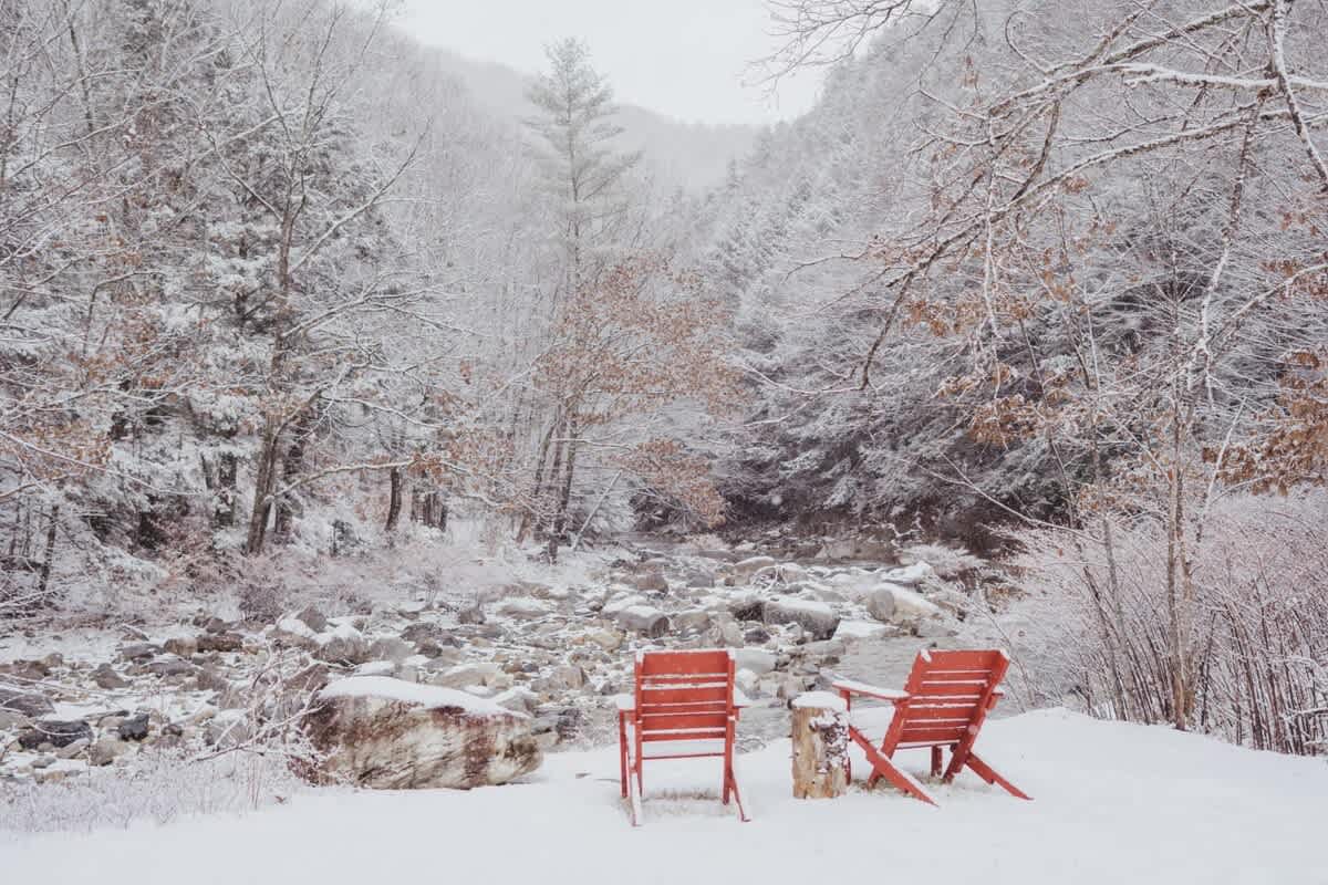 Two vibrant red Adirondack chairs sit in deep snow beside a snow-covered riverbed, surrounded by a serene winter forest. Bare trees and snow-dusted evergreens create a peaceful, secluded atmosphere.