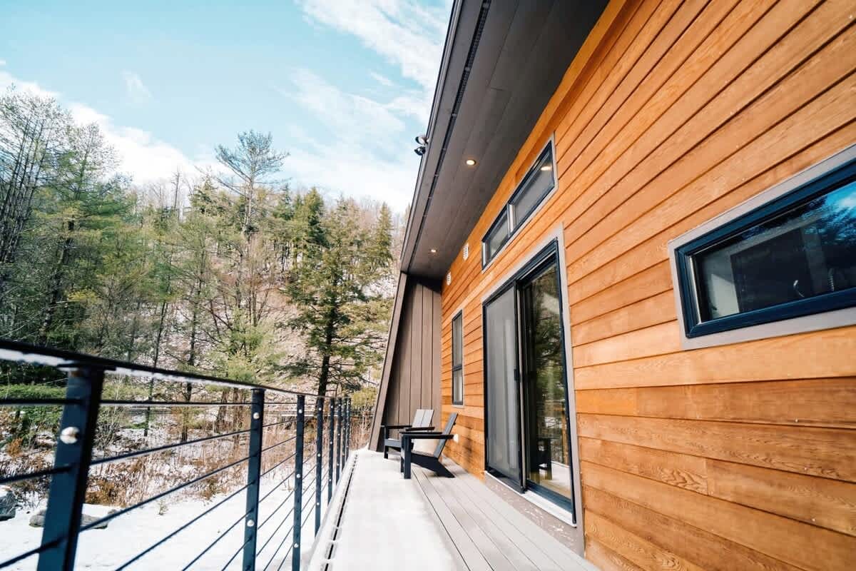 The exterior of a modern house showcases horizontal wood siding and a balcony with a metal cable railing, overlooking a serene snowy, wooded landscape. Two dark chairs are placed on the balcony, inviting outdoor relaxation.