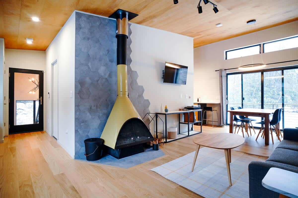 A spacious open-concept living area showcases a distinctive yellow freestanding fireplace set against a hexagonal tile accent wall, with a wall-mounted TV nearby. Hardwood floors extend into the adjacent dining area, creating a cohesive modern space.