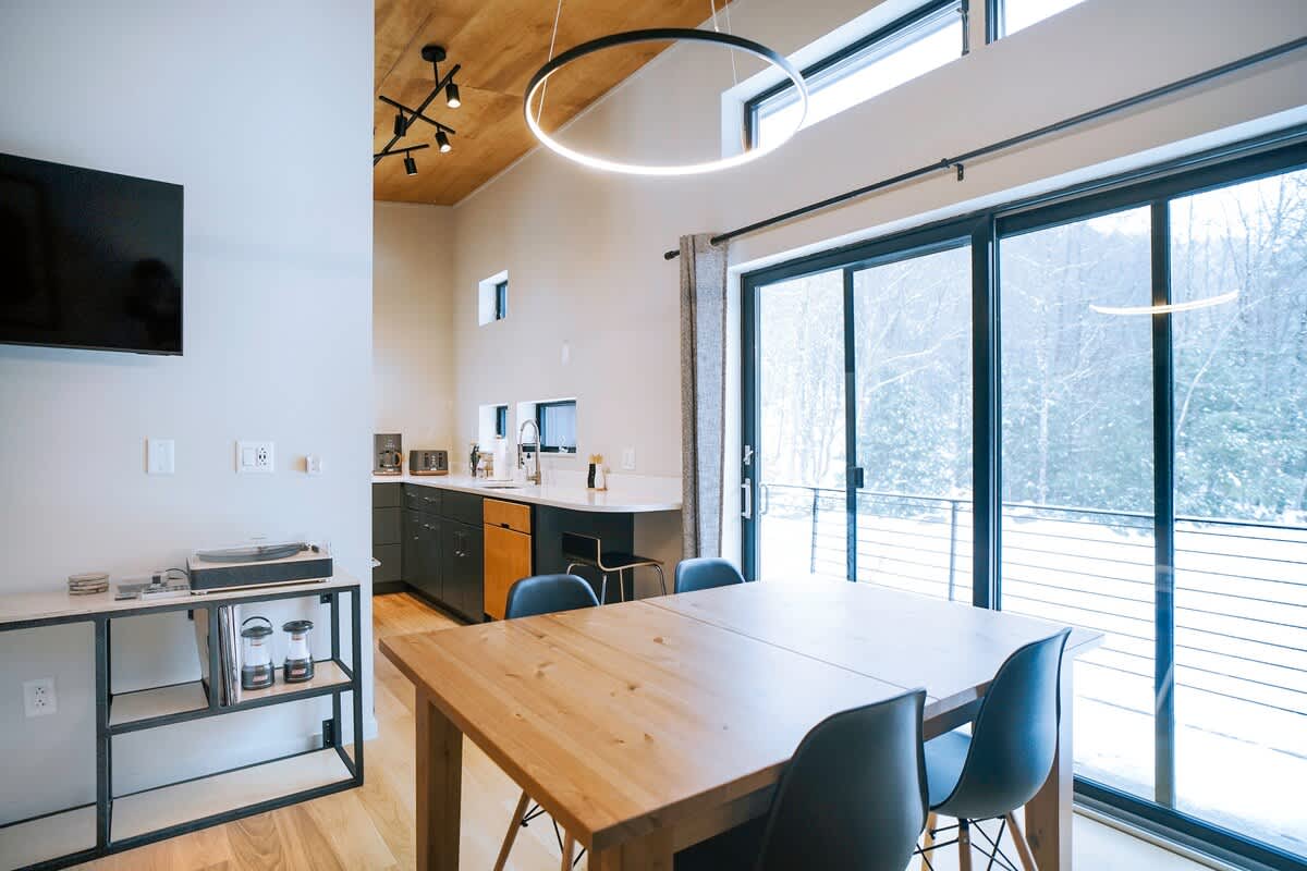 An open-concept dining area features a light wood table with dark chairs, adjacent to a modern kitchen with dark cabinetry and a white island. Large sliding glass doors offer panoramic views of a snowy, wooded landscape, illuminated by a circular LED light fixture.