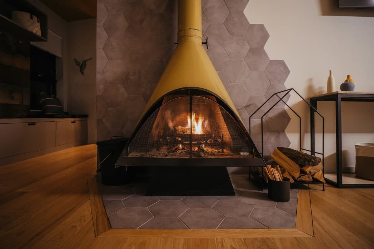 A striking vintage yellow cone-shaped fireplace with a mesh screen, set against a wall with gray hexagonal tiles. A pile of firewood is neatly stacked beside the fireplace, ready for use.