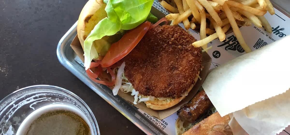 A close-up shot of a crispy fried chicken sandwich with lettuce, tomato, and pickles, served on a metal tray alongside golden french fries and a glass of beer.