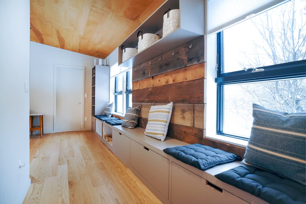 A functional mudroom features a long built-in window seat with storage cabinets and plush cushions, set against a rustic wooden accent wall. Large windows offer views of bare trees, and the space is topped with a warm wood-paneled ceiling.