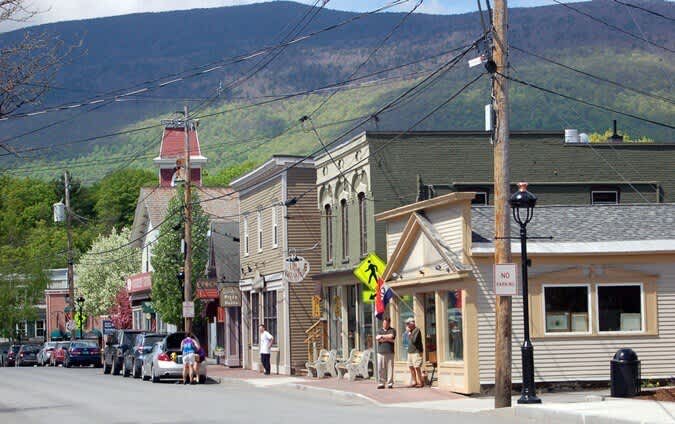 A charming small town street features historic storefronts with awnings and intricate details, parked cars, and pedestrians, all set against a backdrop of lush green mountains under a clear sky.