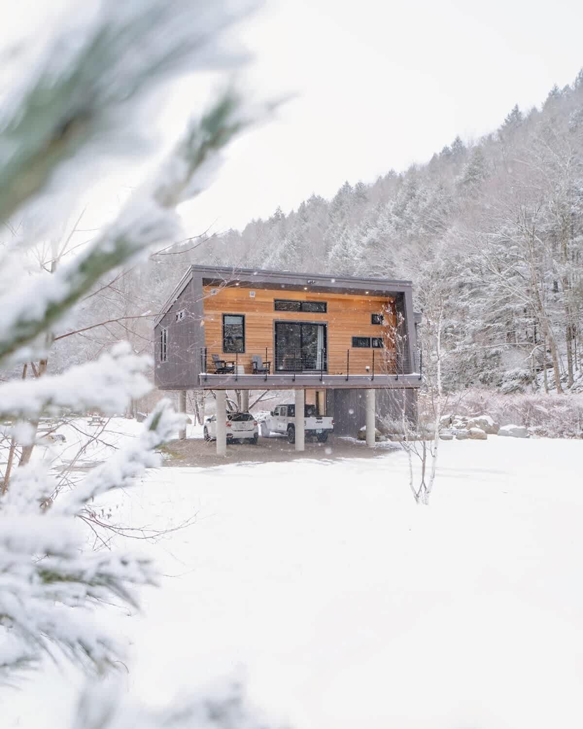 Modern elevated house with wood and dark gray siding, standing out against a pristine snow-covered landscape during winter. Snow-laden trees frame the view, with cars visible parked beneath the elevated structure.